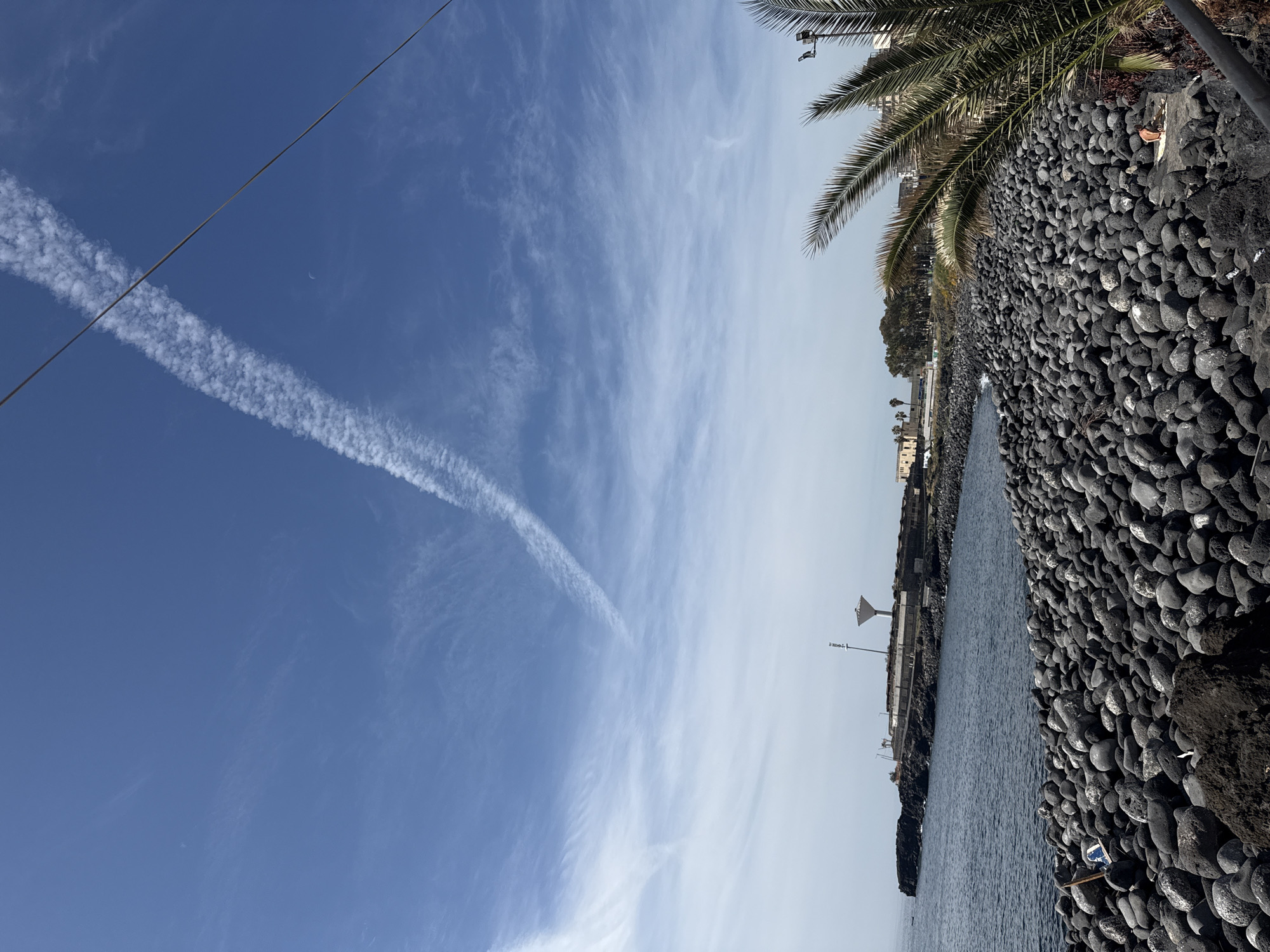 The image depicts a scene in an unknown city. The foreground features a rocky embankment made of dark, rounded stones, bordering a body of water, presumably the sea. A palm tree is partially visible on the right side of the frame, suggesting a warm climate. In the distance, structures and buildings are discernible along the coastline. The sky, occupying a significant portion of the image, is a mix of blue and light clouds, with a prominent contrail bisecting the frame, created by a passing aircraft. A power line is visible in the sky.