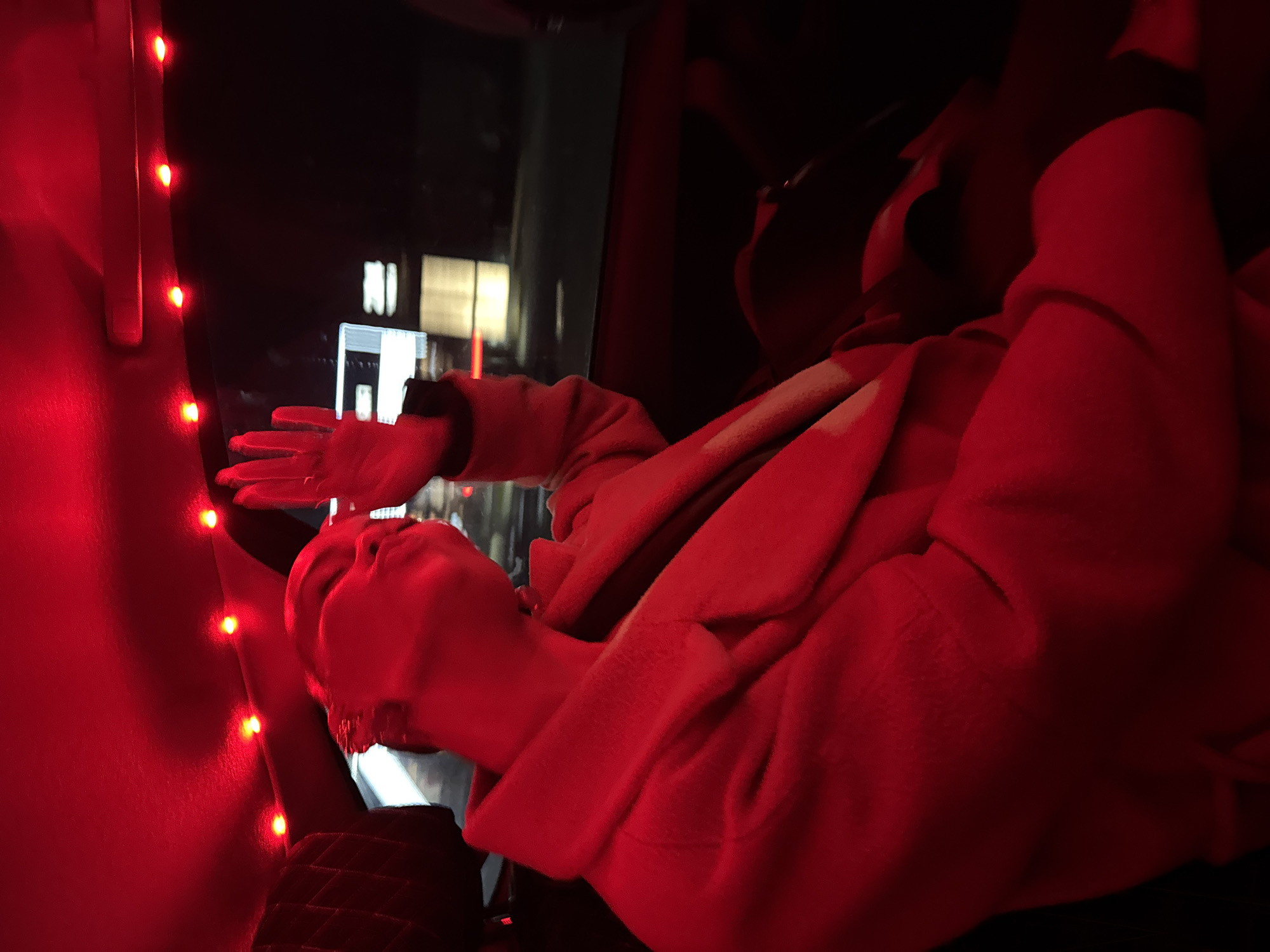 This media file depicts an indoor scene at night, heavily illuminated by a strong red light. The primary subject is a person, possibly in Chaikovsky, Russia, seen from a side-back perspective, wearing a warm, reddish-orange coat or jacket. Their head is tilted slightly back as they look outwards, and their right hand is extended towards a vertical string of glowing red LED lights, which are fixed along a dark interior surface, likely a window frame or wall. A watch is visible on their right wrist.
Through the window, blurred reflections and distant lights of buildings are discernible against the dark night sky, confirming the time of day. The interior setting, with its distinct window structure and the arrangement of the lights, suggests it could be within a vehicle such as a bus or train, or a room with a large window. Another figure or part of a person is faintly visible in the dark background, further inside. The predominant red lighting creates a warm, atmospheric, and intimate mood. The person appears to be engaged in a moment of calm observation or contemplation. There is no visible text in the image.