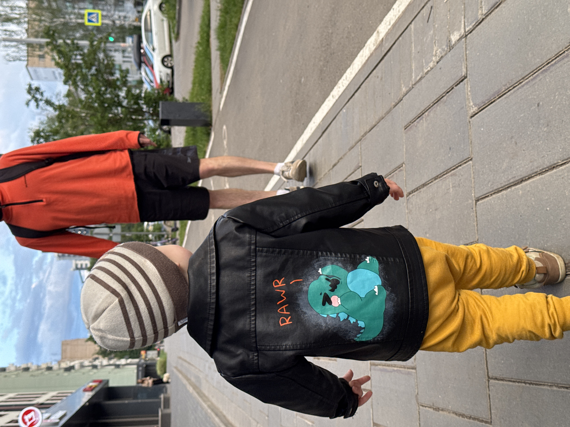 A low-angle, medium shot depicts a young child walking on a sidewalk next to a street. The child is wearing a striped beanie hat, a black leather jacket with a cartoon dinosaur and "RAWR!" painted on the back, and yellow pants. The child is facing away from the camera, walking toward the right. An adult wearing an orange jacket and black shorts is walking behind the child. The sidewalk is paved with grey rectangular stones. A street runs parallel to the sidewalk, with a white car and some greenery visible on the far side. Buildings are visible in the background. A sign with the number 1 on it is visible on the left side of the picture. The location is Izhevsk, Russia.