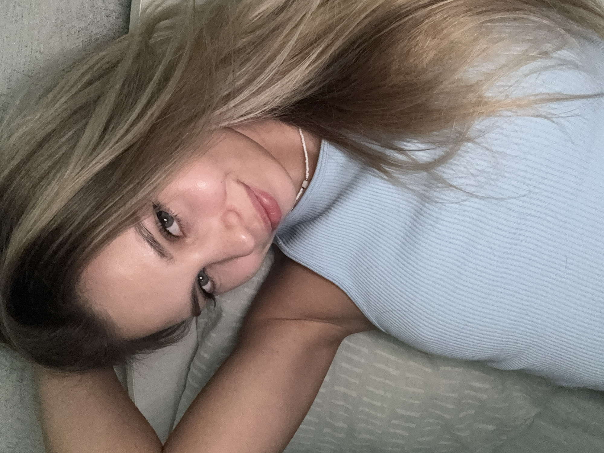 The image is a close-up, selfie-style shot of a young woman with long, blonde hair. She is wearing a light blue ribbed tank top and a delicate white beaded necklace. Her left arm is raised, with her hand behind her head. She is looking directly at the camera with blue eyes and a slight smile. The background appears to be a light-colored, textured surface, possibly a wall or bedding. The lighting suggests an indoor setting, likely at night or in a dimly lit room. There is no identifiable location in the city of Chaikovsky, Russia.