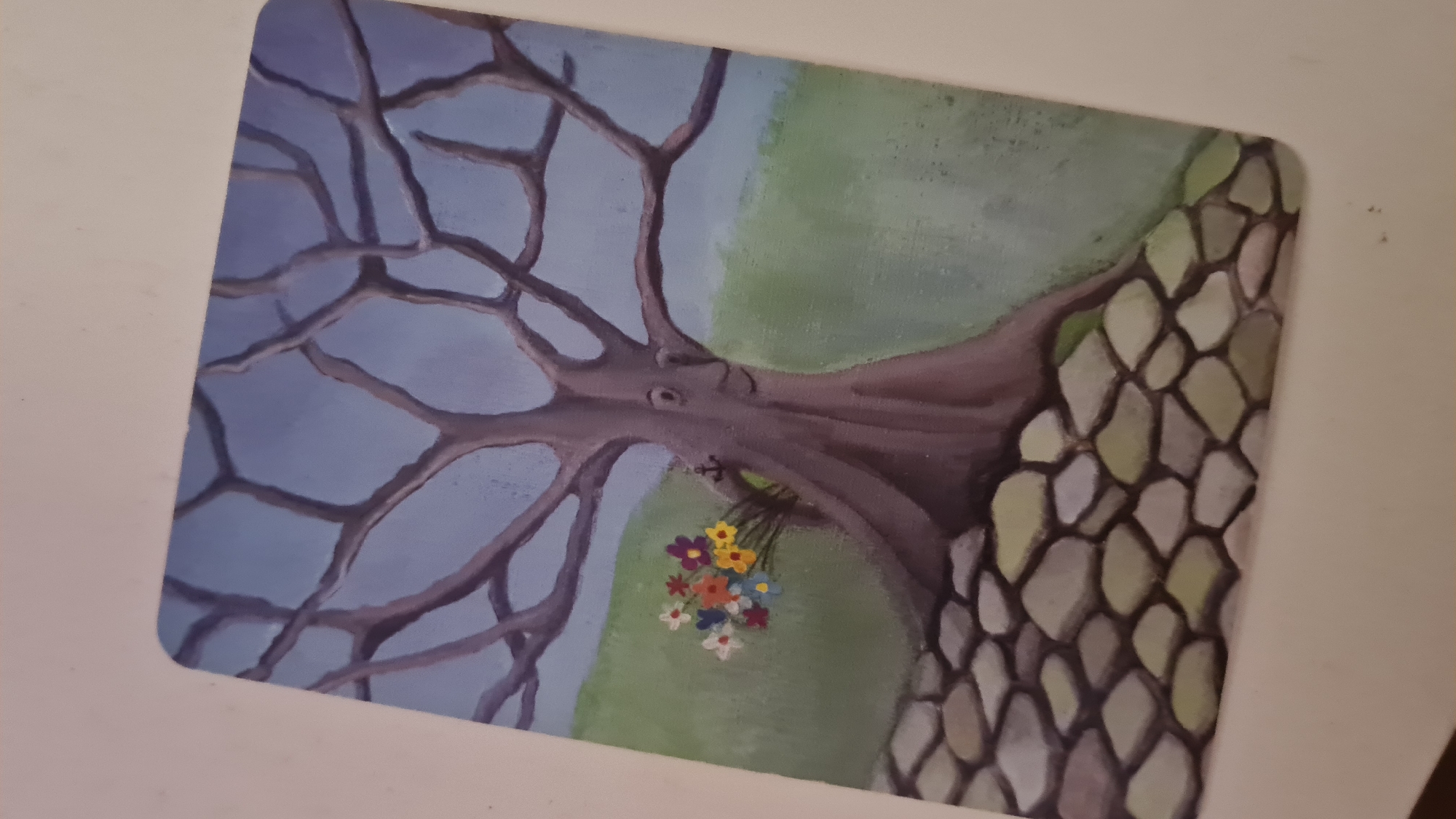The image depicts a painting or print of a stylized tree with bare branches against a blue sky. A patch of green grass or foliage is visible behind the tree. A stone wall is depicted on the right side of the artwork. A small bouquet of colorful flowers hangs from one of the branches. The location of the scene, as specified, is in the city of Lachish Regional Council, Israel.