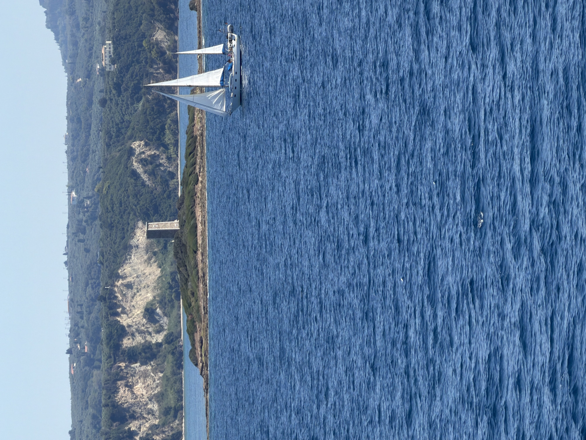 The image captures a sunlit coastal scene in the Municipal Unit of Kastro - Kyllini, Greece. In the foreground, a vast expanse of deep blue water is textured with gentle ripples. Further out, a white sailboat with its sails filled navigates the sea. In the middle ground, a small, rocky island is visible, topped with a stone tower. The background features a densely forested hillside, characteristic of the Mediterranean landscape, with scattered buildings and the faint outline of mountains under a clear blue sky. The overall impression is one of tranquility and natural beauty.