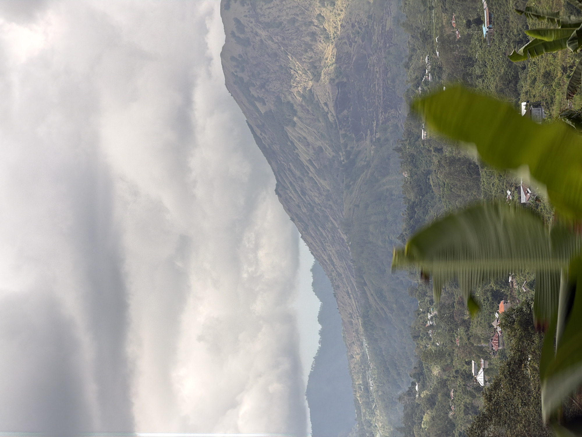 The image depicts a panoramic view of a mountainous landscape near Kintamani, Indonesia, under a cloudy sky. In the foreground, large, blurred green banana leaves partially obscure the view. Beyond them, a dense, green, and hilly terrain is dotted with scattered houses and buildings, some with red roofs. The middle ground features a vast, sloping mountain range covered in vegetation and marked by visible geological features, possibly lava flows from past volcanic activity. The background is dominated by a large, prominent mountain with a textured, barren upper slope, indicative of volcanic ash and rock. The overall environment suggests a rural or semi-rural setting within a volcanic region.