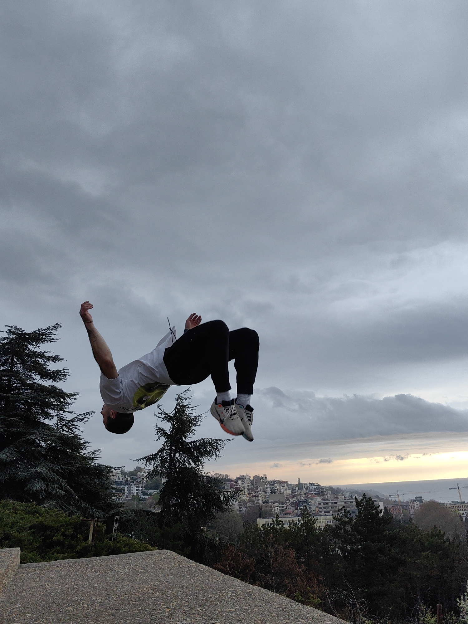 A man is performing a backflip in front of a cityscape. He is wearing a white t-shirt, black pants, and sneakers. The city is identified as Varna, Bulgaria, as indicated by the buildings and the presence of the Black Sea in the background. The location appears to be an elevated park area, with trees visible in the foreground and a concrete structure upon which the man is presumably standing. The sky is overcast with a sunset in the distance over the sea.