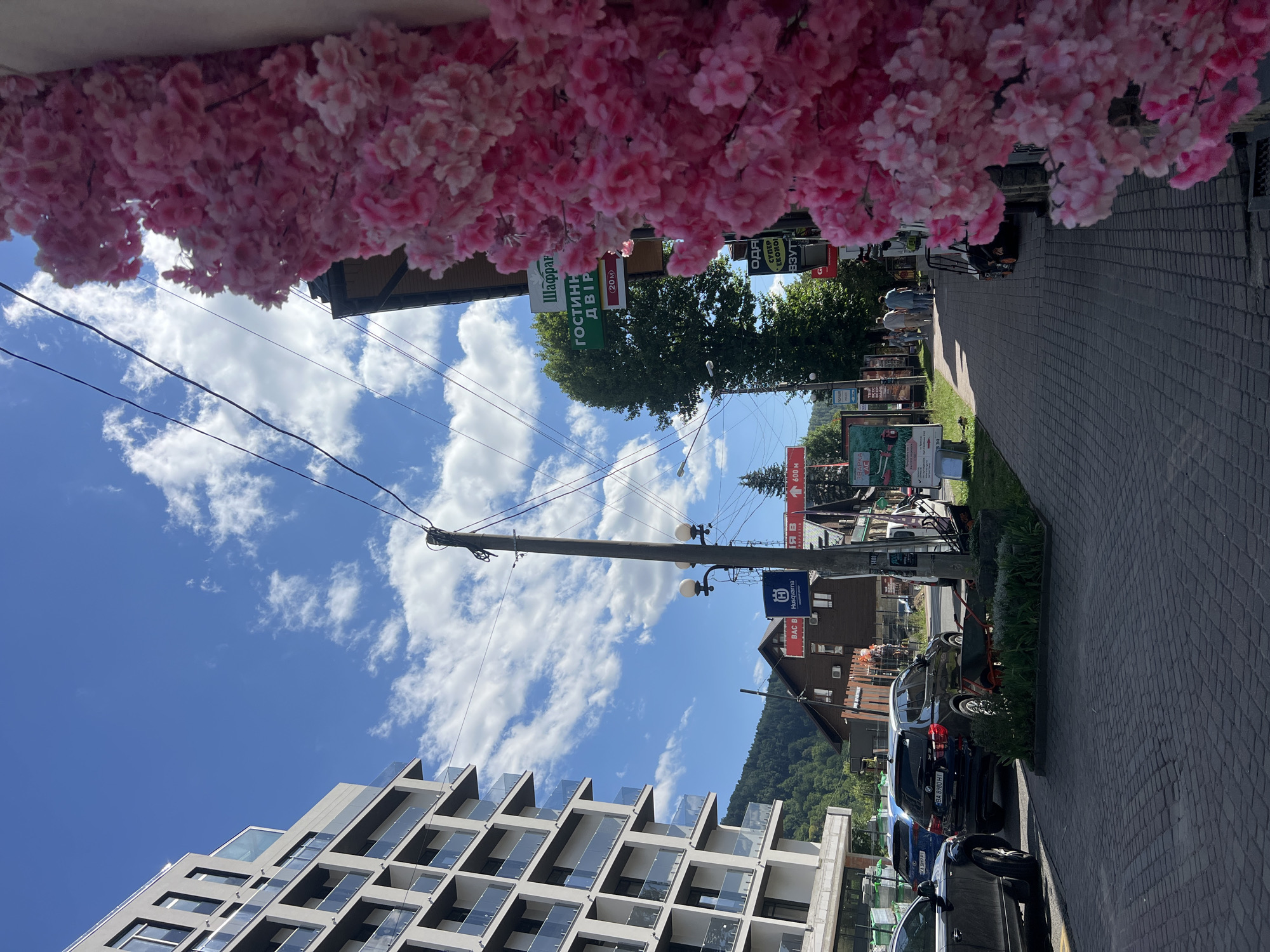 A street scene in Yaremche, Ukraine, features a modern multi-story building on the left, a paved sidewalk with several cars parked along the street on the right. A bright pink artificial floral arrangement is prominently displayed on the right side of the frame. Multiple signs are visible, including one that reads "ГОСТИННИЙ ДВІР" (Guest Courtyard) and another with the Husqvarna logo. A few people are walking on the sidewalk in the distance, and lush green trees and hills form the backdrop under a blue sky with scattered clouds. The sun is shining, casting shadows on the pavement.