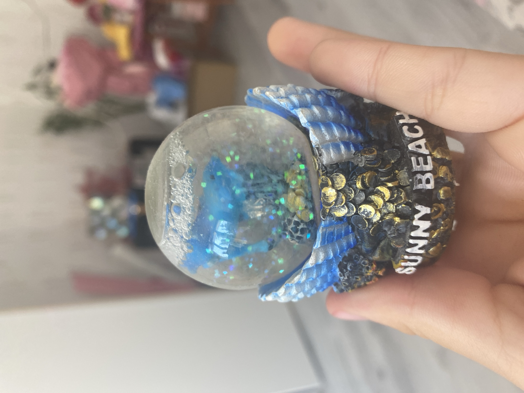 A person's hand holds a snow globe that features a marine scene. Inside the globe, blue glitter and small sea creatures are visible, suspended in liquid. The base of the snow globe is decorated with what appears to be scales or shells in blue and white, and gold accents with the words "SUNNY BEACH" inscribed in white lettering. The background is blurred, showing a domestic indoor environment with furniture and a pink bouquet. The image was taken in Blagoevgrad, Bulgaria.
