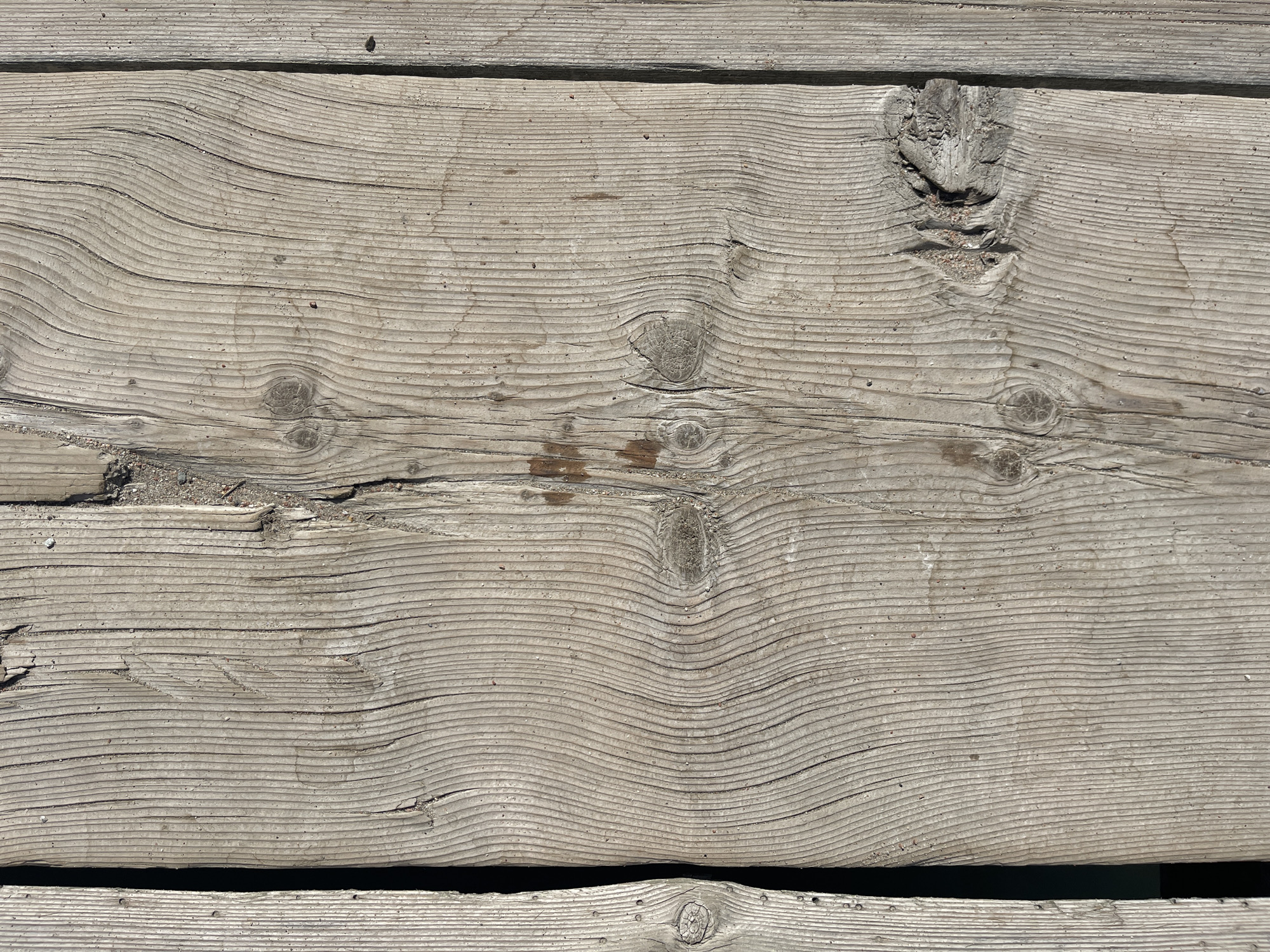 The image shows a close-up view of weathered wooden planks. The wood grain is prominent, with vertical lines running along the length of the planks. There are several knots visible, some of which are partially filled with dirt or sand. The wood appears to be rough and aged, suggesting it may be part of an outdoor structure. The lighting creates shadows that highlight the texture of the wood. There are no people or specific identifiable landmarks present in the image. The location is described as Ornek, Kyrgyzstan, though no visual cues in the image confirm this.