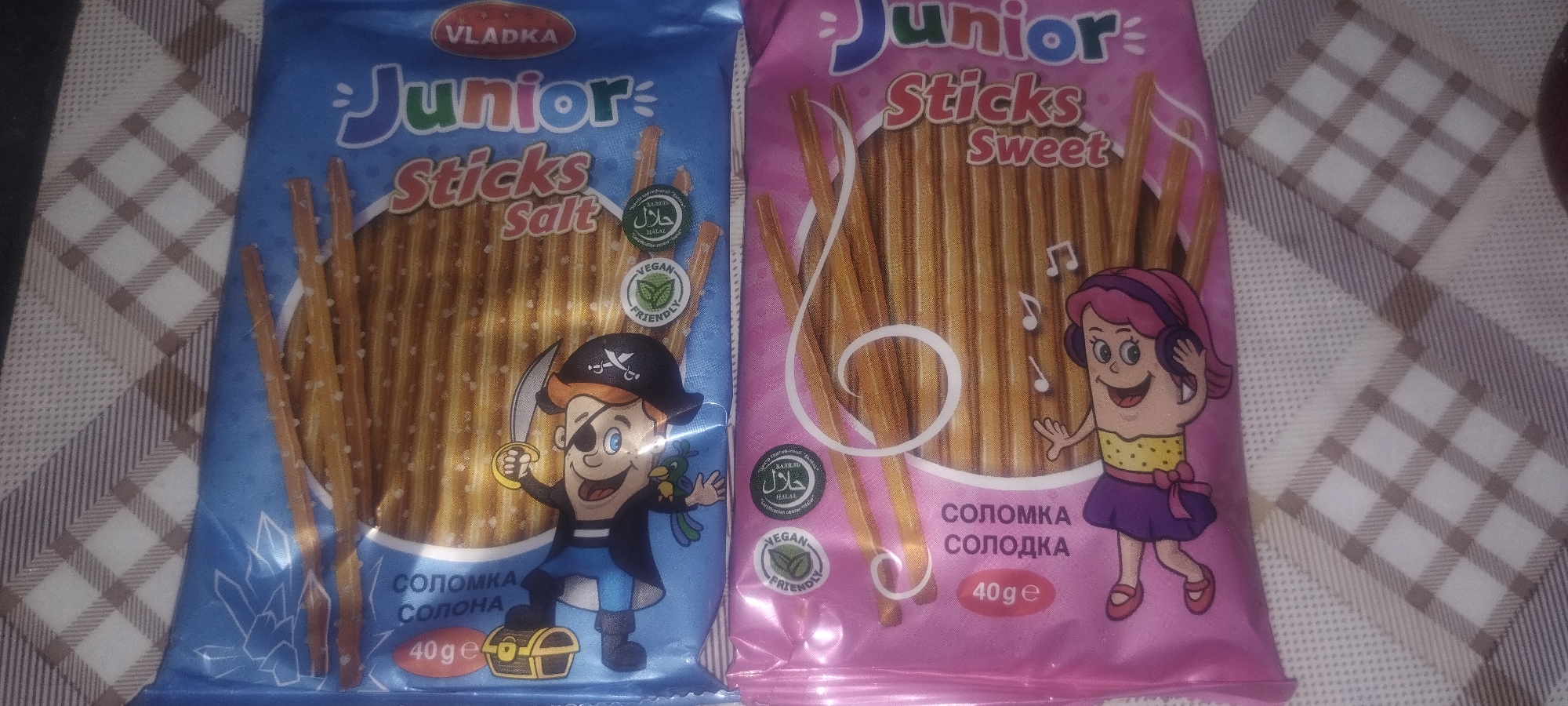 Two packages of snack sticks are shown on a checkered surface. The package on the left is blue and labeled "VLADKA Junior Sticks Salt". It features a cartoon pirate character and a parrot. The package on the right is pink and labeled "Junior Sticks Sweet". It depicts a cartoon girl with headphones and musical notes. Both packages show stacks of slender, golden-brown snack sticks. Nutritional and certification labels, including "HALAL" and "VEGAN FRIENDLY," are visible on both packages. The location is identified as Unknown, Unknown.