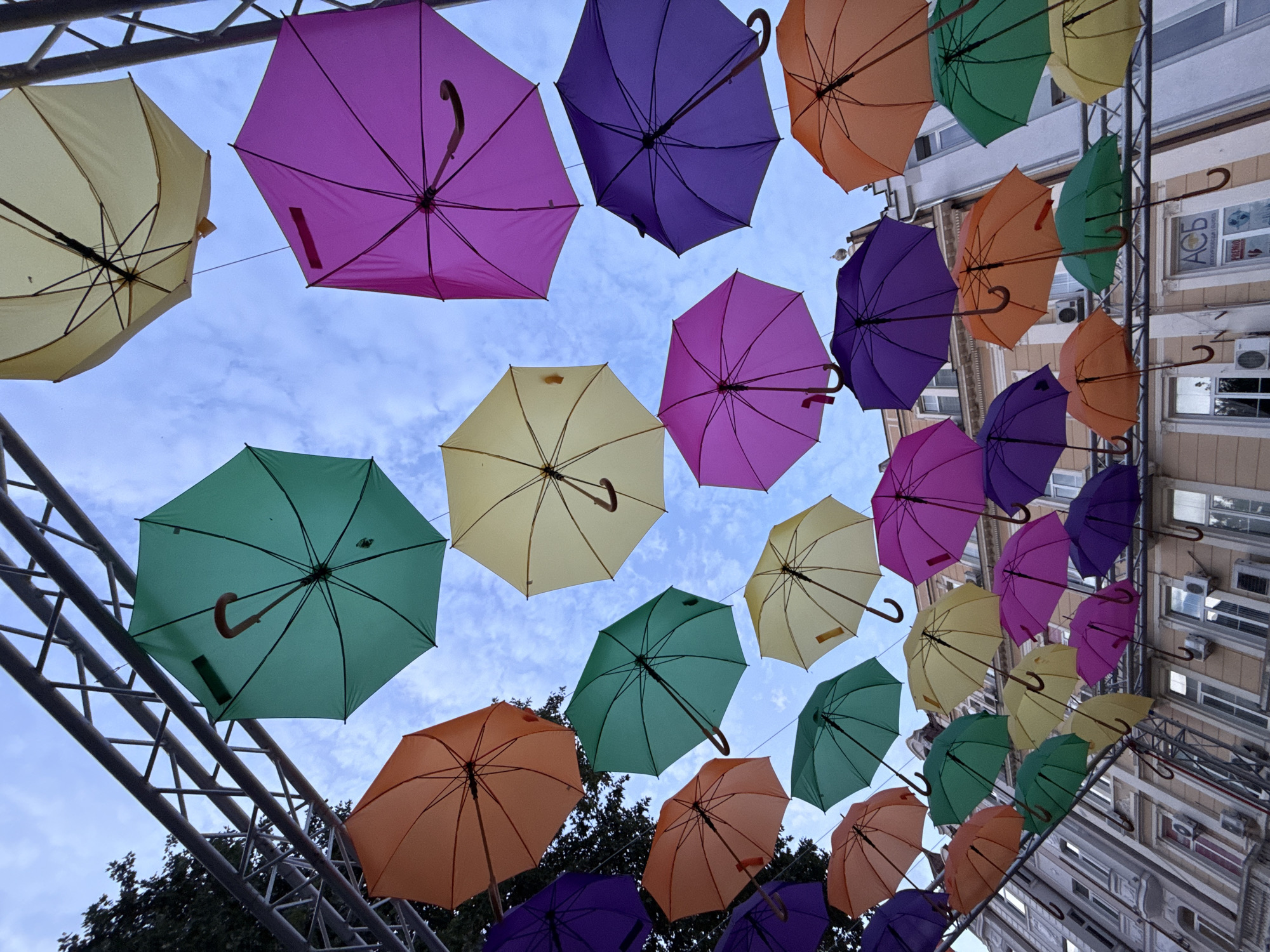 The image captures a vibrant street art installation in Plovdiv, Bulgaria, featuring a dense arrangement of colorful umbrellas suspended overhead against a cloudy sky. The umbrellas, in shades of yellow, green, purple, and orange, are affixed to metal scaffolding that spans across a street. Below the umbrellas, the upper facades of old, ornate buildings are visible, characterized by their traditional architecture, large windows, and visible air conditioning units. The scene is devoid of people, creating a sense of quiet observation of the art.