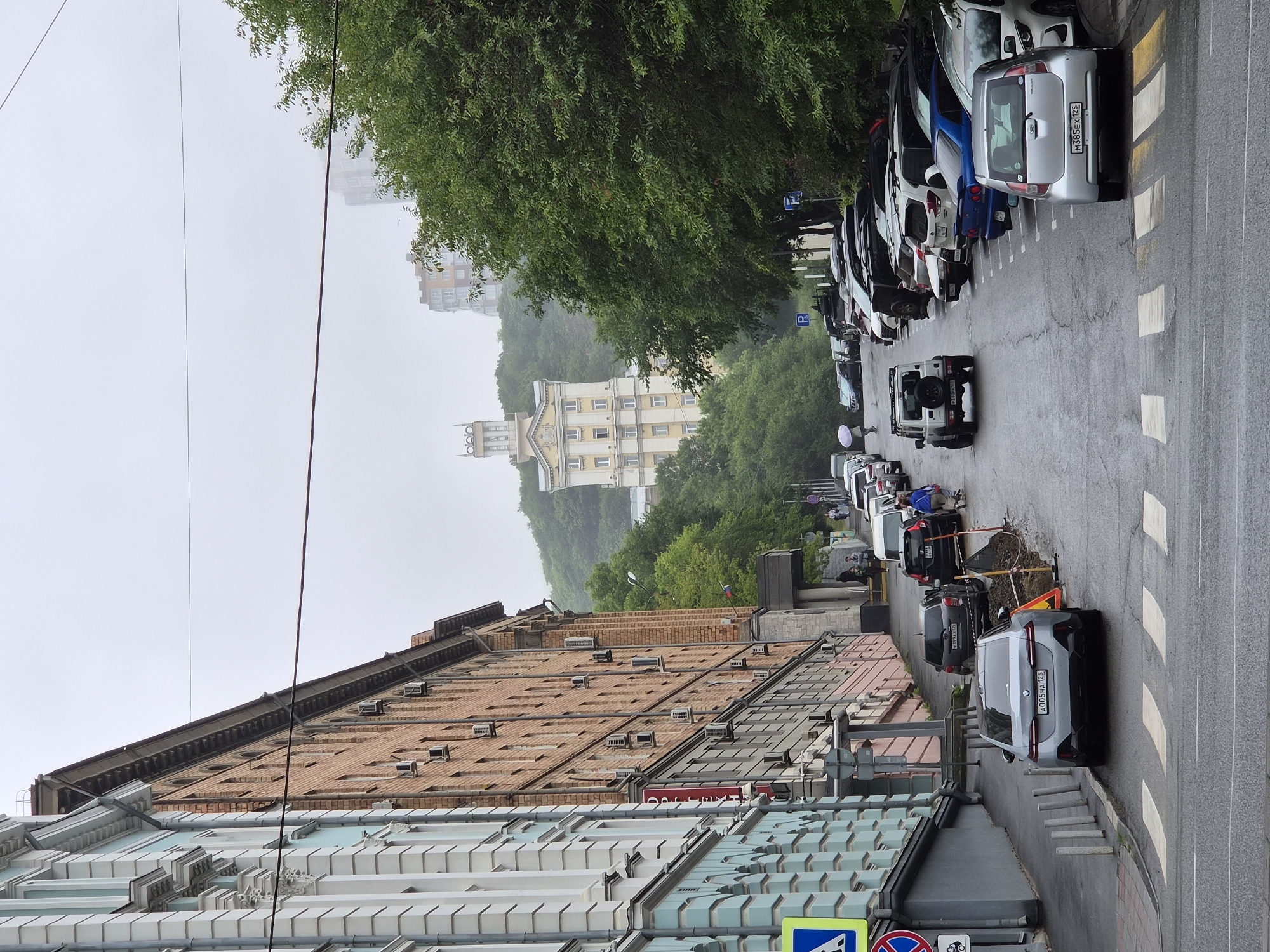 The image depicts a street scene in Vladivostok, Russia, likely on a cloudy or foggy day. On the left, a multi-story building with a light blue facade and ornate white detailing dominates the foreground. Adjacent to it is a brick building with numerous windows and air conditioning units. The street slopes downwards, with parked cars lining both sides. Several pedestrians are visible, some walking and others standing.
In the background, a distinctive yellow and white building with a prominent tower stands on a tree-covered hill. Another modern high-rise building is visible further in the distance, partially obscured by mist. Trees and greenery line the right side of the street. Traffic signs and road markings, including a pedestrian crossing, are present. Construction or repair work is evident on the side of the road in the midground, marked by orange cones and barriers. The overall impression is of an urban environment with a mix of older and newer architecture, set against a hilly landscape.