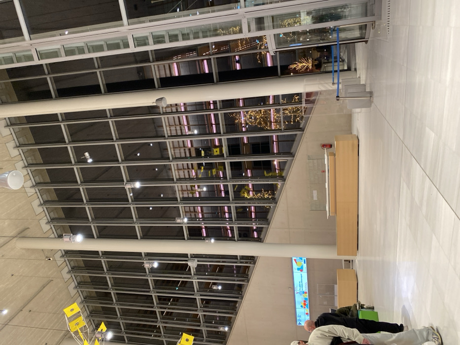The image displays the interior of a large, modern public building, likely a lobby or atrium, located in Kallithea, Greece. The dominant feature is a high, sloped ceiling constructed from a grid of glass and metal panels, allowing views of the exterior or an adjacent area.
The building's interior is well-lit by numerous small, recessed lights within the ceiling structure and from cylindrical fixtures. Reflections of warm-toned decorative lights, possibly from outdoor trees or festive installations, are prominent in the glass panels, suggesting it is evening or nighttime.
In the mid-ground, a long, light-wood reception desk or counter is situated against a plain wall. Clear plexiglass barriers are installed on the counter, and a red fire extinguisher is mounted on the wall behind it. To the right of the desk, blue stanchion ropes are visible, indicating a queue or restricted access point, possibly near an entrance.
In the lower right corner, several individuals are partially visible, appearing to be waiting or resting. One person wears a white jacket, another a dark one. On the left side, yellow, kite-like decorative elements are suspended from the ceiling structure.
A brightly lit digital display or poster board, partially visible in the lower-center, features colorful graphics. Some text is discernable, including "You're all set to go!". The overall scene conveys a spacious, contemporary, and functional environment.