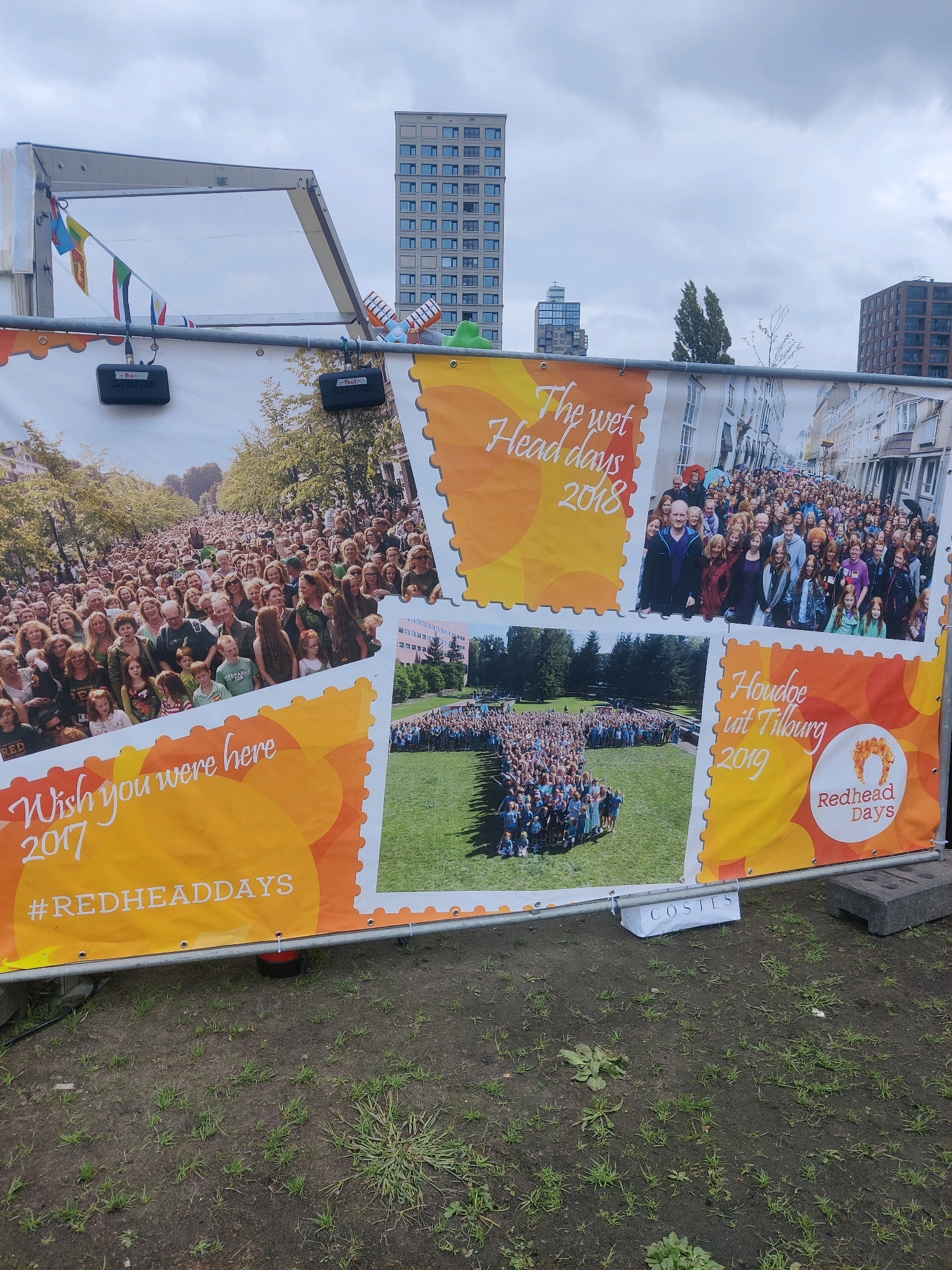 The image is a banner displayed outdoors, likely in Tilburg, Netherlands, given the text "Houdoe uit Tilburg 2019" which translates to "Goodbye from Tilburg 2019." The banner is made of a large, horizontal fabric print. It is supported by a metal frame and appears to be for an event called "Redhead Days."
The banner features three distinct photographic sections, each framed to resemble a postage stamp, with a torn edge effect.
The top left section shows a crowd of people, predominantly with red hair, gathered outdoors on a tree-lined street. The text "Wish you were here 2017 #REDHEADDAYS" is overlaid on this section in a stylized font.
The top right section displays a large group of people, again with a significant number having red hair, standing in a street lined with buildings. The text "The wet Head days 2018" is superimposed on this image.
The bottom left section features a high-angle aerial view of a large gathering of people arranged in the shape of a heart on a grassy field.
The bottom right section contains a circular logo with a stylized image of red hair in the center, along with the text "Redhead Days." Below this, the text "Houdoe uit Tilburg 2019" is visible.
The banner is set against a cloudy sky and appears to be situated on uneven, grassy ground with some patches of dirt. In the background, several tall modern buildings are visible, including one that is distinctly tall and light-colored with many windows. There are also some smaller buildings and trees. Two black audio speakers are mounted on the metal support structure of the banner. A white sign with the word "COSTES" is placed on the ground near the bottom right of the banner.