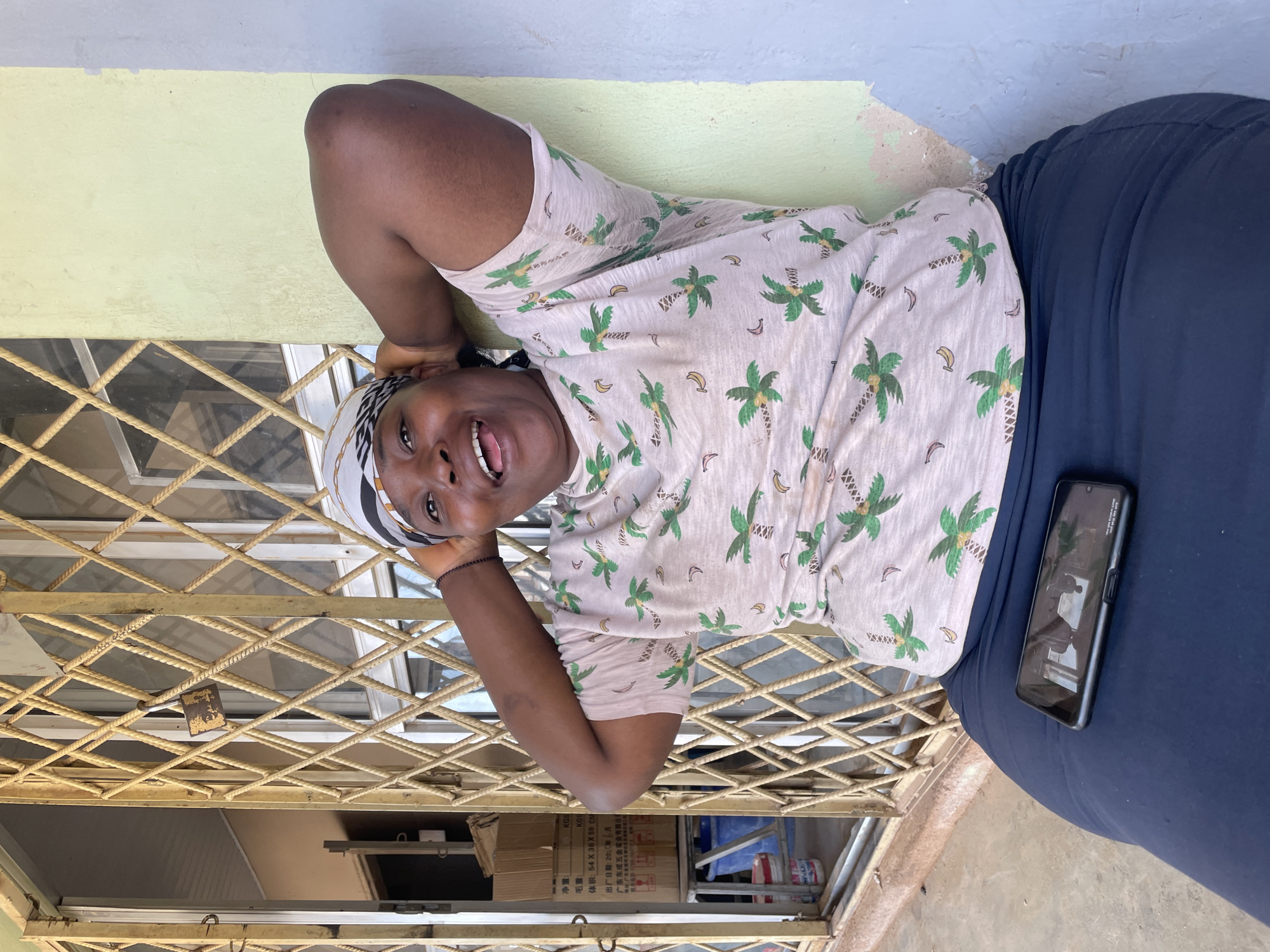 A woman is depicted outdoors, leaning against a light green wall and a metal gate in Barakin Akawo, Nigeria. She is wearing a light-colored t-shirt with a palm tree and banana pattern and a headscarf. Her arms are behind her head as she smiles at the camera. A dark blue skirt covers her lower body, and a smartphone rests on her lap. Behind her, the metal gate is closed, and beyond that, a window is visible. Boxes and buckets are stacked behind the gate. The ground is paved. The lighting suggests it is daytime.