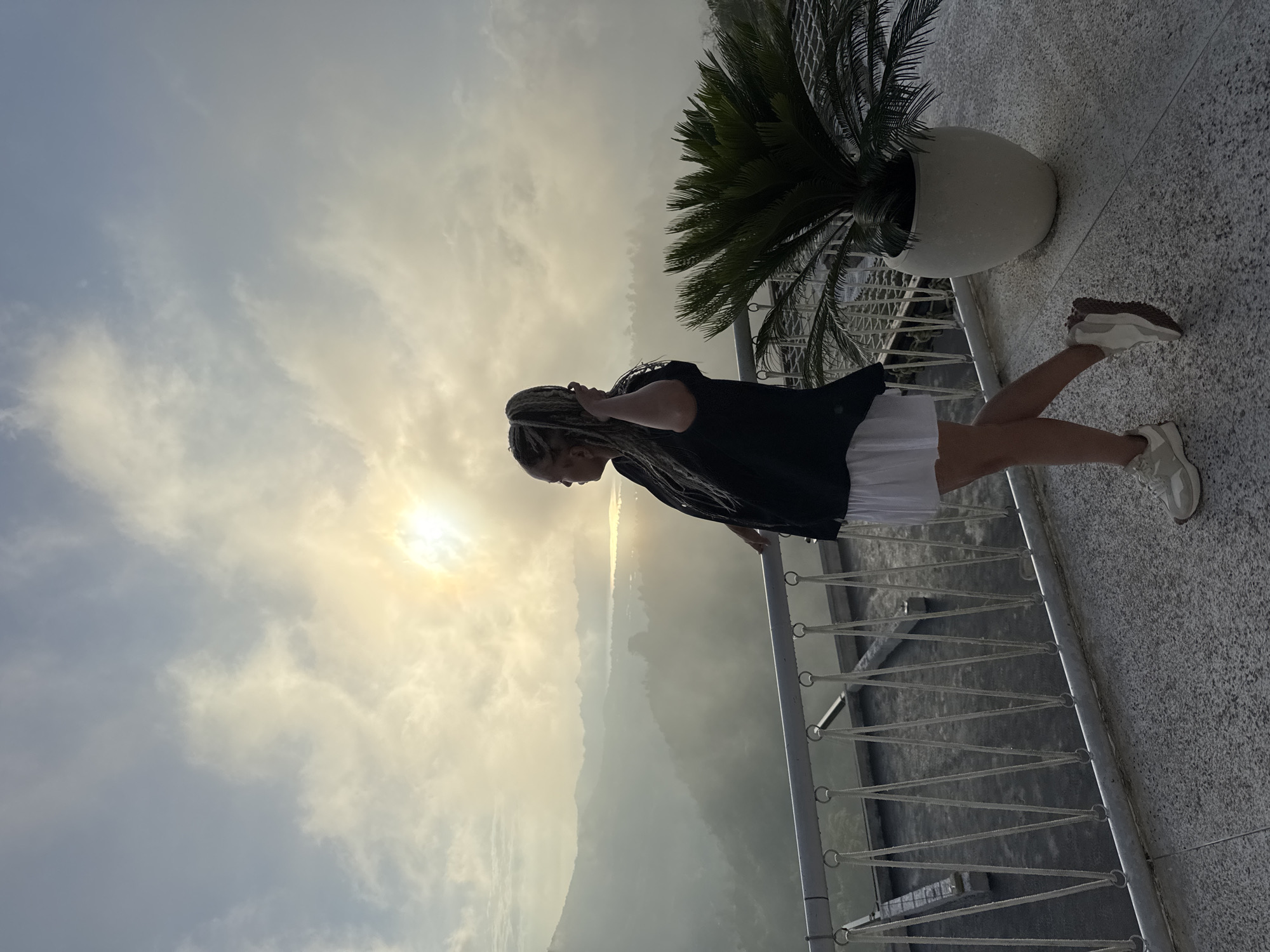A woman with blonde braids stands on a balcony overlooking a mountainous landscape. The sun shines brightly in the sky, obscured by some clouds. The balcony has a metal railing and is decorated with a large potted plant with lush green fronds. The woman wears a black shirt, white skirt, and white shoes. The location is Kedisan, Indonesia.