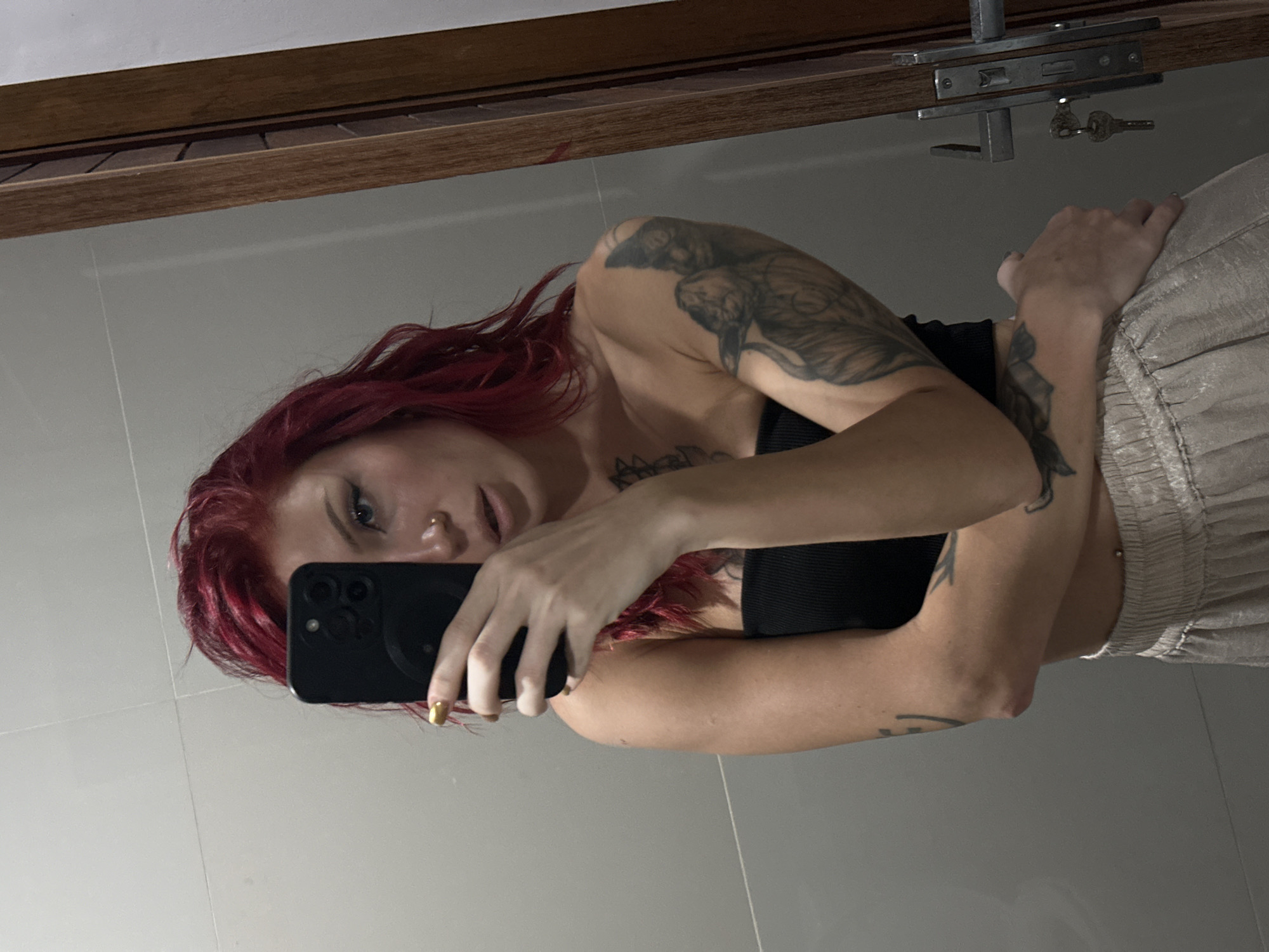 A young woman with striking vibrant red hair is captured in a vertical mirror selfie. She holds a black smartphone, likely an iPhone due to its distinctive triple-camera module, in her right hand, with her metallic gold-painted fingernails visible. The phone covers part of her face, leaving her blue eyes and a septum nose piercing visible. She is wearing a dark, possibly black, bandeau-style top that reveals her shoulders and midriff. Her arms are extensively covered with detailed tattoos. She also wears light-colored, shimmering pants with a gathered or elastic waistband, and a small piercing is visible on her side above the pants line.
The background suggests an indoor setting, possibly a bathroom or dressing area, with light-colored, likely tiled, walls. Above her reflection, a wooden structure, appearing to be a frame or a shelf, spans the upper portion of the scene. Attached to this wooden element is an older metal locking mechanism, from which two traditional keys hang. The woman looks directly at the phone’s camera, conveying a neutral expression. The image was taken in North Kuta, Indonesia. No discernible text is visible within the image.
