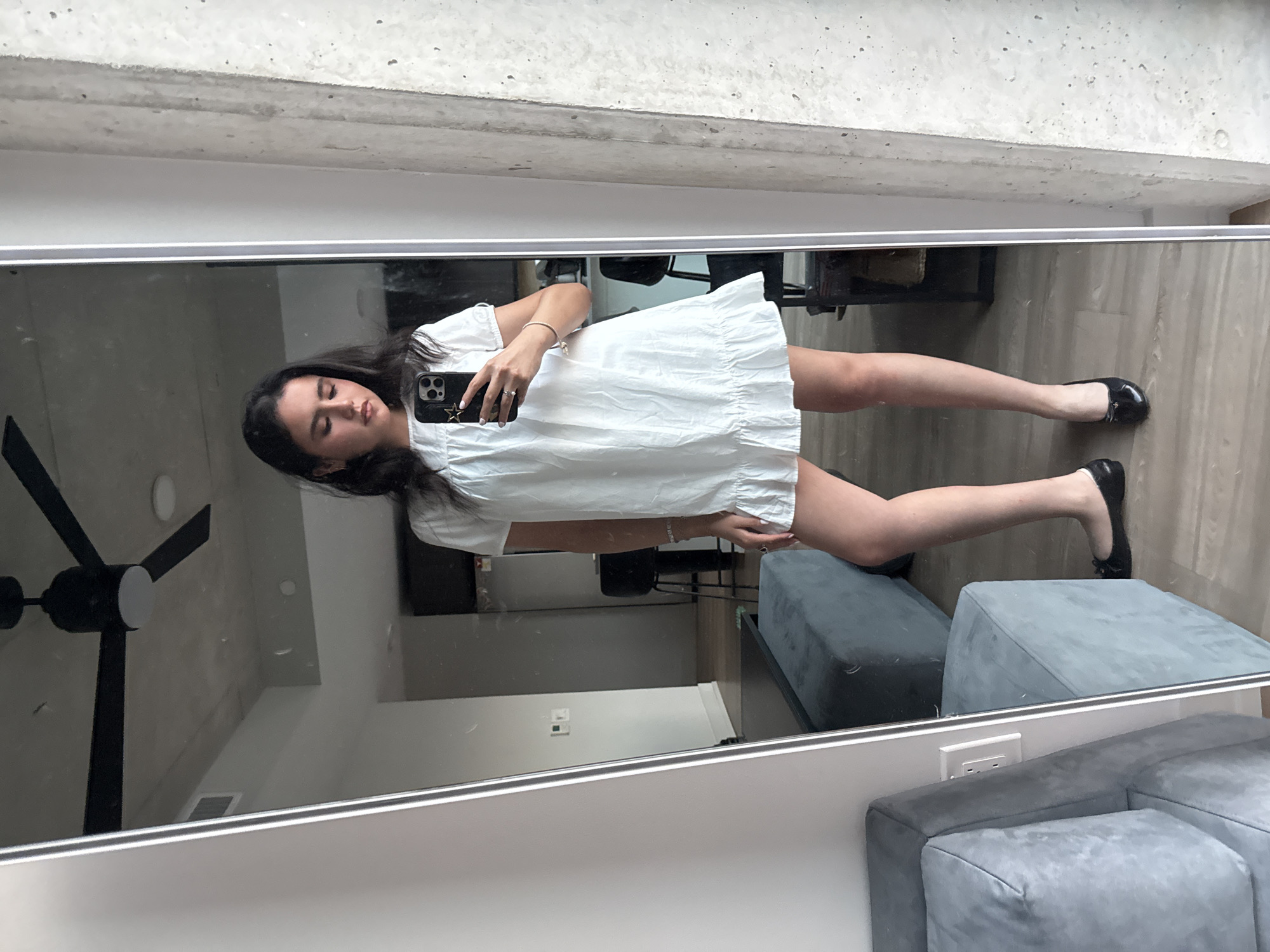 A young woman stands in front of a floor-to-ceiling mirror, taking a selfie with her smartphone. She is wearing a white, short-sleeved dress with a ruffled hem and black ballet flats. The mirror reflects her and the surrounding apartment, which features a ceiling fan, a gray sectional sofa, and a concrete accent wall. The woman is holding her phone in her right hand, and her left arm rests at her side. She appears to be indoors, likely in a residential setting in Austin, Texas. The lighting is natural, coming from an unseen window.
