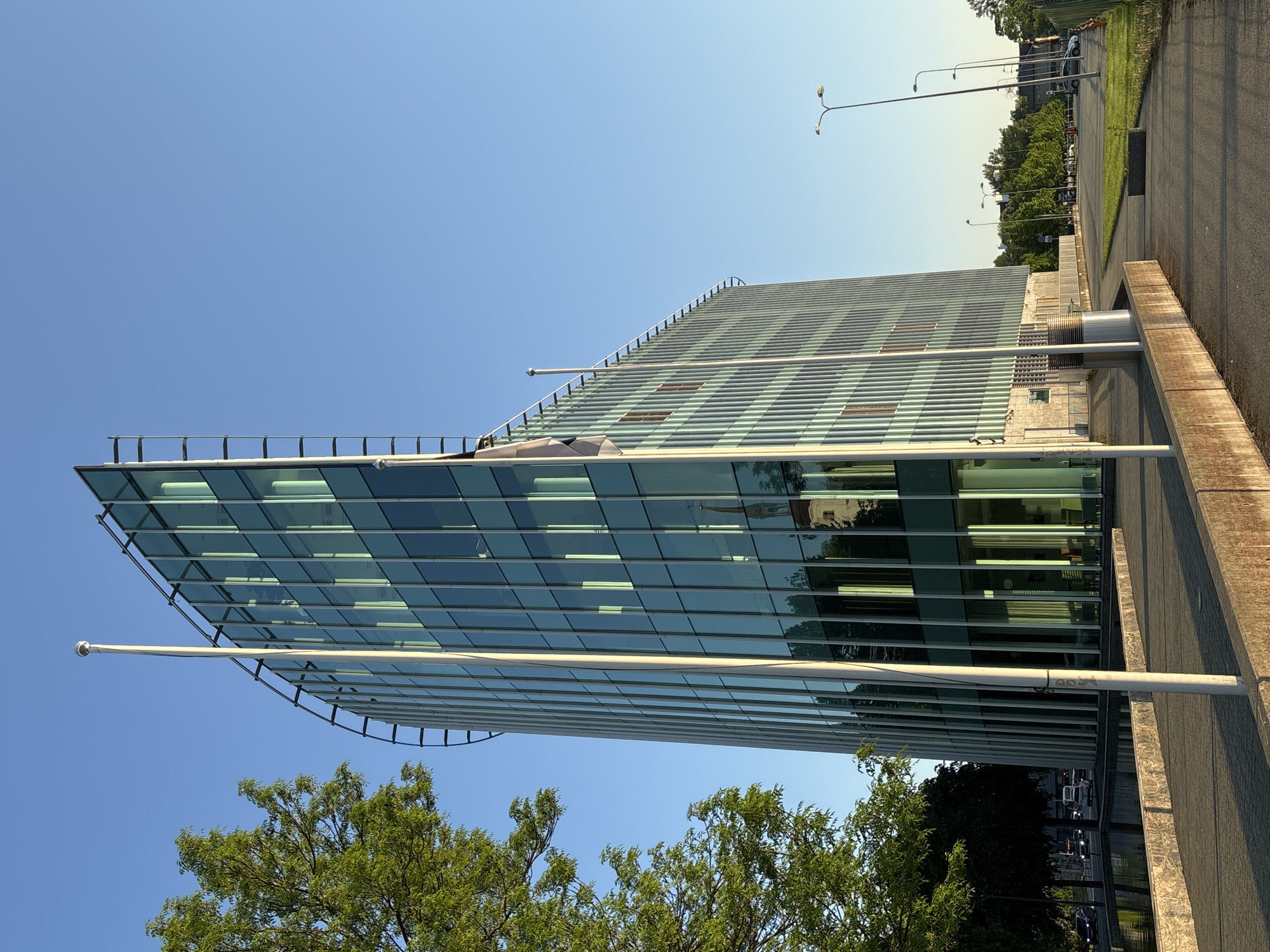 This image captures a modern, multi-story building under a clear, bright blue sky, likely in Tallinn, Estonia. The building features a distinctive contemporary architectural style, characterized by extensive use of glass and metal. One prominent face of the building, which appears to be its front or top section, has a striking sloped or curved profile, entirely clad in reflective glass panels that mirror the sky and surrounding greenery. The other visible side of the structure also features glass panels, some of which are overlaid with horizontal metallic louvers or bands, adding texture to the facade.
In the foreground, two tall, white flagpoles stand upright, extending beyond the frame towards the top, with their tips visible near the building's edge. Lush green trees are visible in the lower left foreground, and more trees line a road in the background, partially obscuring distant vehicles. Along the paved area next to the building, several modern streetlights with multiple lamp heads are visible, illuminating the scene. A low concrete or stone barrier runs parallel to the building on the right side of the paved area.
The overall impression is one of clear, sunny daylight, likely morning or afternoon, given the crisp shadows and vibrant light. No specific activity or event is taking place, presenting a static view of the urban architecture. There is no discernible text visible in the image.
