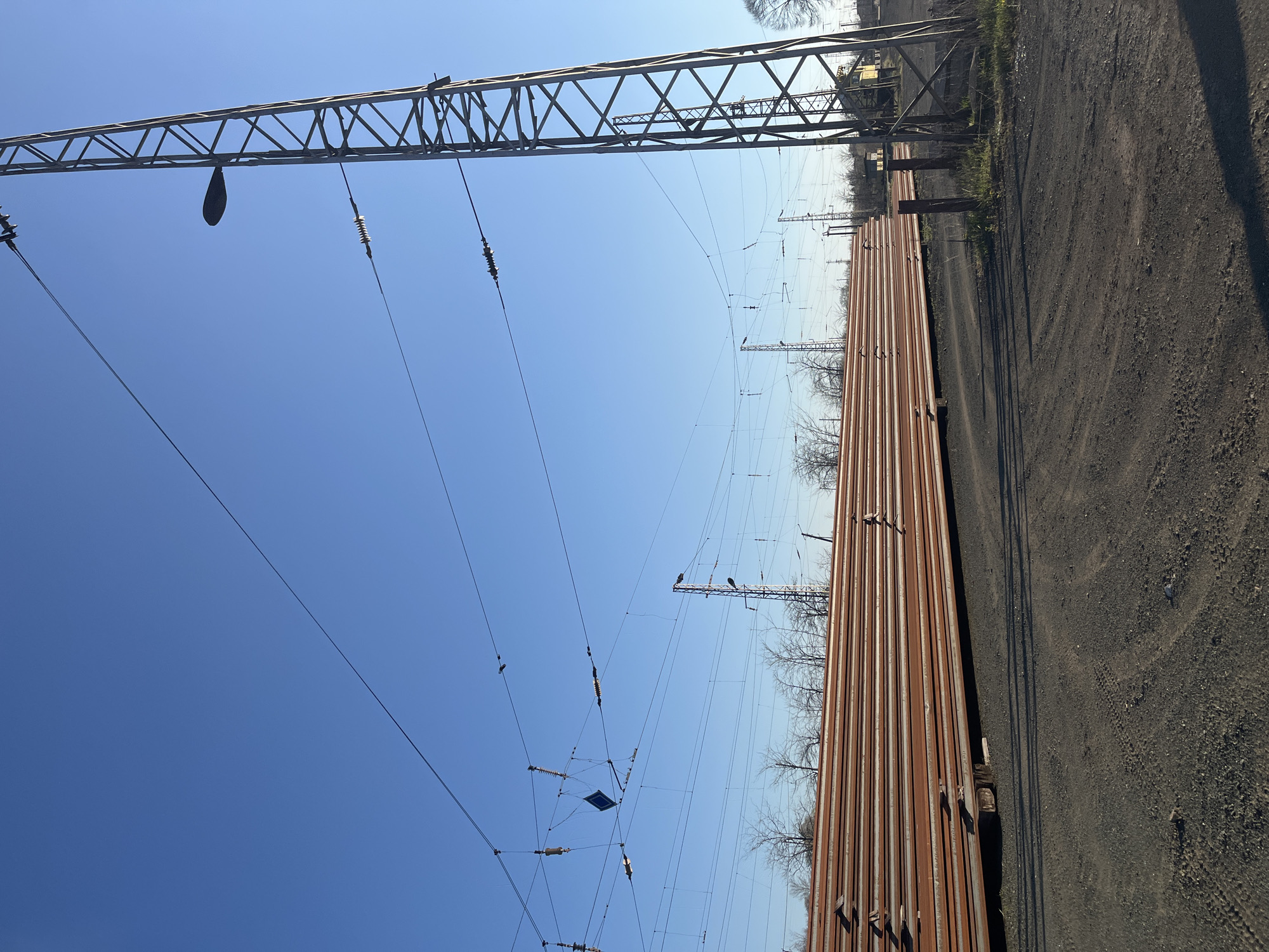 The image shows a sunny outdoor scene in Lozarevo, Bulgaria, near a railway. A metal lattice electrical tower stands to the left, supporting high-voltage power lines. Several steel rails are stacked in parallel rows on the ground. Beyond the rails is bare earth. Bare branches of trees are visible in the background. The sky is clear and blue.