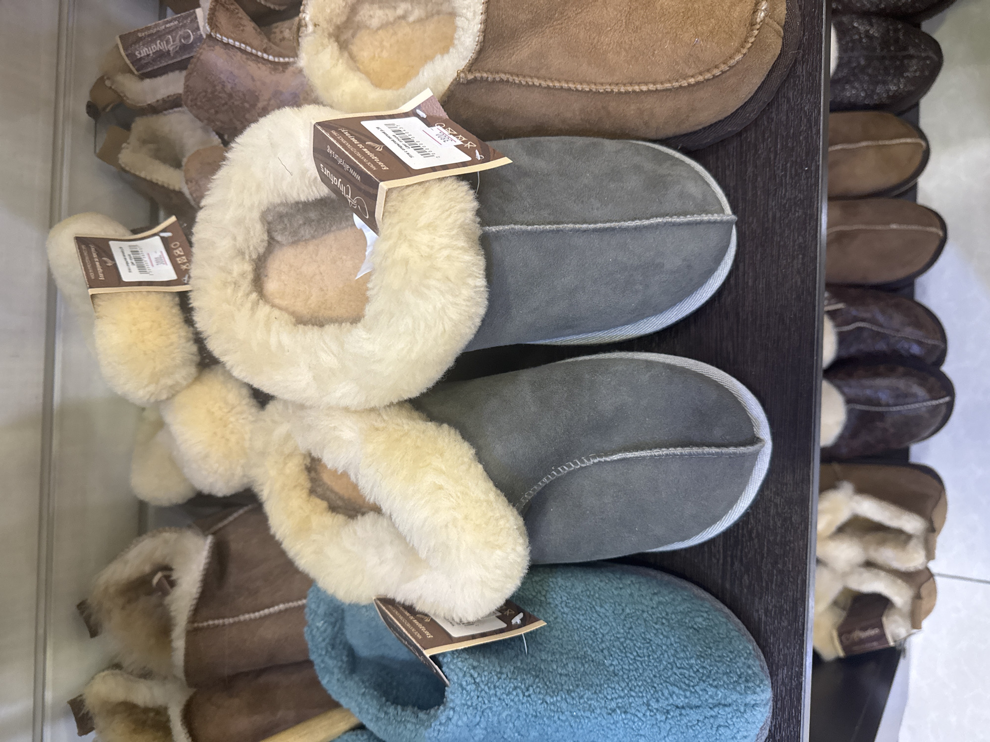 A close-up, eye-level shot shows a retail display of sheepskin slippers on a dark shelf. The slippers are lined with fluffy white shearling. In the foreground, from left to right, are a teal slipper, a grey slipper, and a brown slipper. A second row of slippers is partially visible below the shelf. Two of the slippers in the front row have tags with the brand name "Alyaturs" and Cyrillic text, indicating they are likely sold in Bishkek, Kyrgyzstan. The lighting is bright, highlighting the textures of the sheepskin.