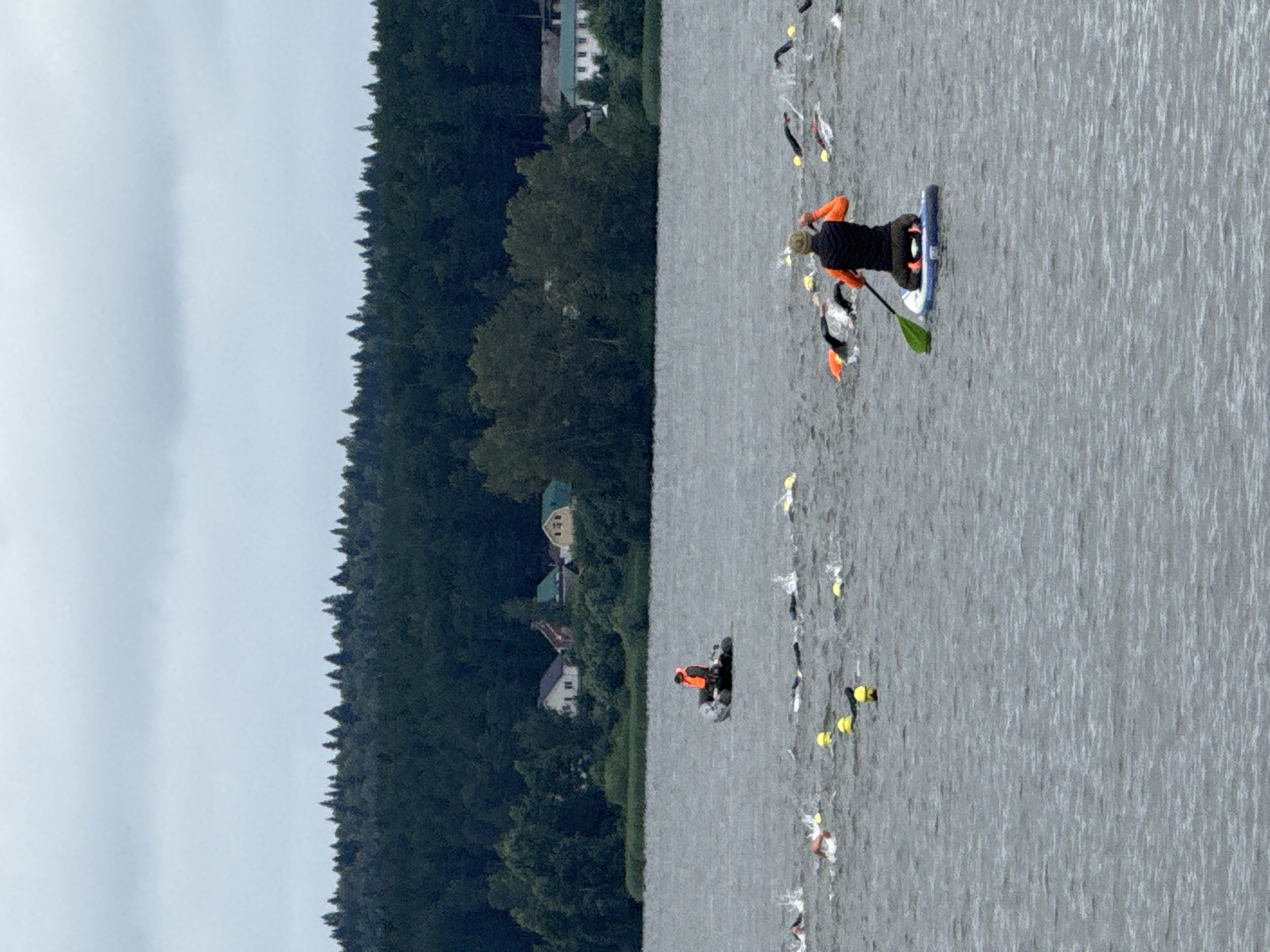 The image depicts an outdoor swimming event taking place on a large body of water, likely a lake or reservoir, under an overcast sky. In the foreground, a person is kneeling on a stand-up paddleboard, holding a paddle. This individual is wearing an orange vest over a dark sleeveless jacket and dark pants. Further out on the water, a group of swimmers is visible, many wearing brightly colored swim caps, some with buoys or floats alongside them. In the mid-ground, a small inflatable boat with a person in an orange vest is present, possibly acting as a safety or support vessel. The background features a dense forest line with scattered buildings visible among the trees, suggesting a semi-rural or suburban setting. The water is rippled, indicating some wind activity. The overall scene suggests a competitive or recreational swimming race. While the specific location within Votkinsk, Russia, cannot be definitively identified from the image alone, the presence of a large body of water and forested surroundings is consistent with the geography of Votkinsk, which is situated on the Kama River and has several lakes.