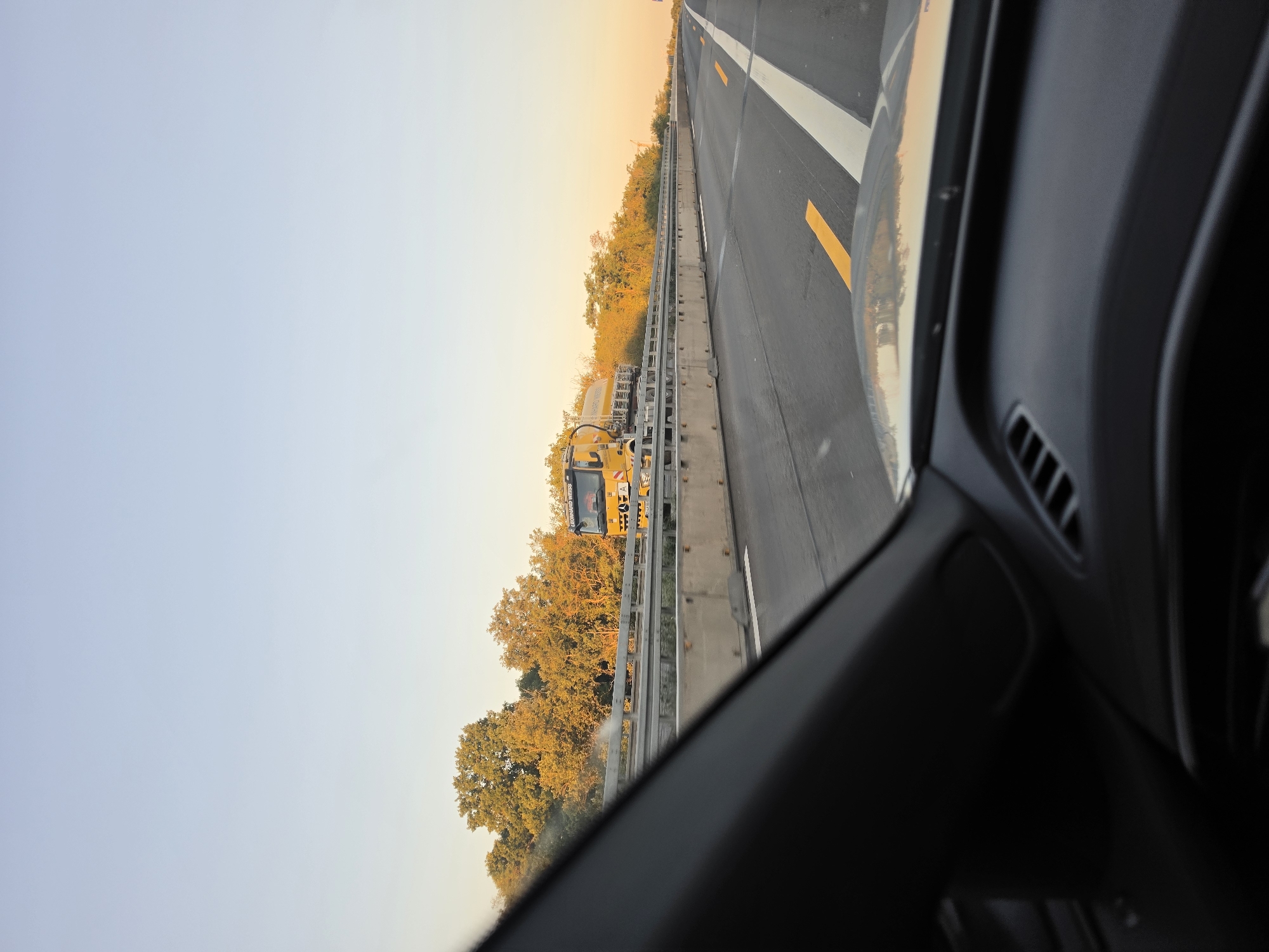 The image, taken from the perspective of a vehicle traveling on a highway, shows a yellow truck with a cylindrical tank on its rear moving in the same direction. The truck has "RENNARD WEISS" written on its side. The scene is illuminated by the warm, low light of sunset, casting long shadows and highlighting the golden hues of the trees lining the highway. The highway itself is asphalt with clear lane markings, including white and yellow lines. A concrete barrier separates the highway lanes from the adjacent foliage. The foreground is dominated by the interior of the vehicle, specifically the dashboard and windshield frame, which are dark and out of focus. The sky is a pale blue, transitioning to a lighter shade near the horizon. The location is identified as Bischofsheim, Germany.