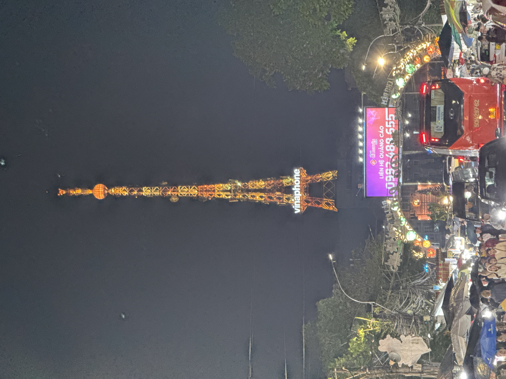 At nighttime, a large transmission tower is lit up and can be seen in the background. At ground level, there are large crowds of people near streets. There is a sign advertising Vinaphone and a large, pink advertising billboard, along with a decorated archway. A red delivery truck is parked on the street. The image appears to have been taken in the city of Da Lat, Vietnam, given the Vinaphone advertisement.
