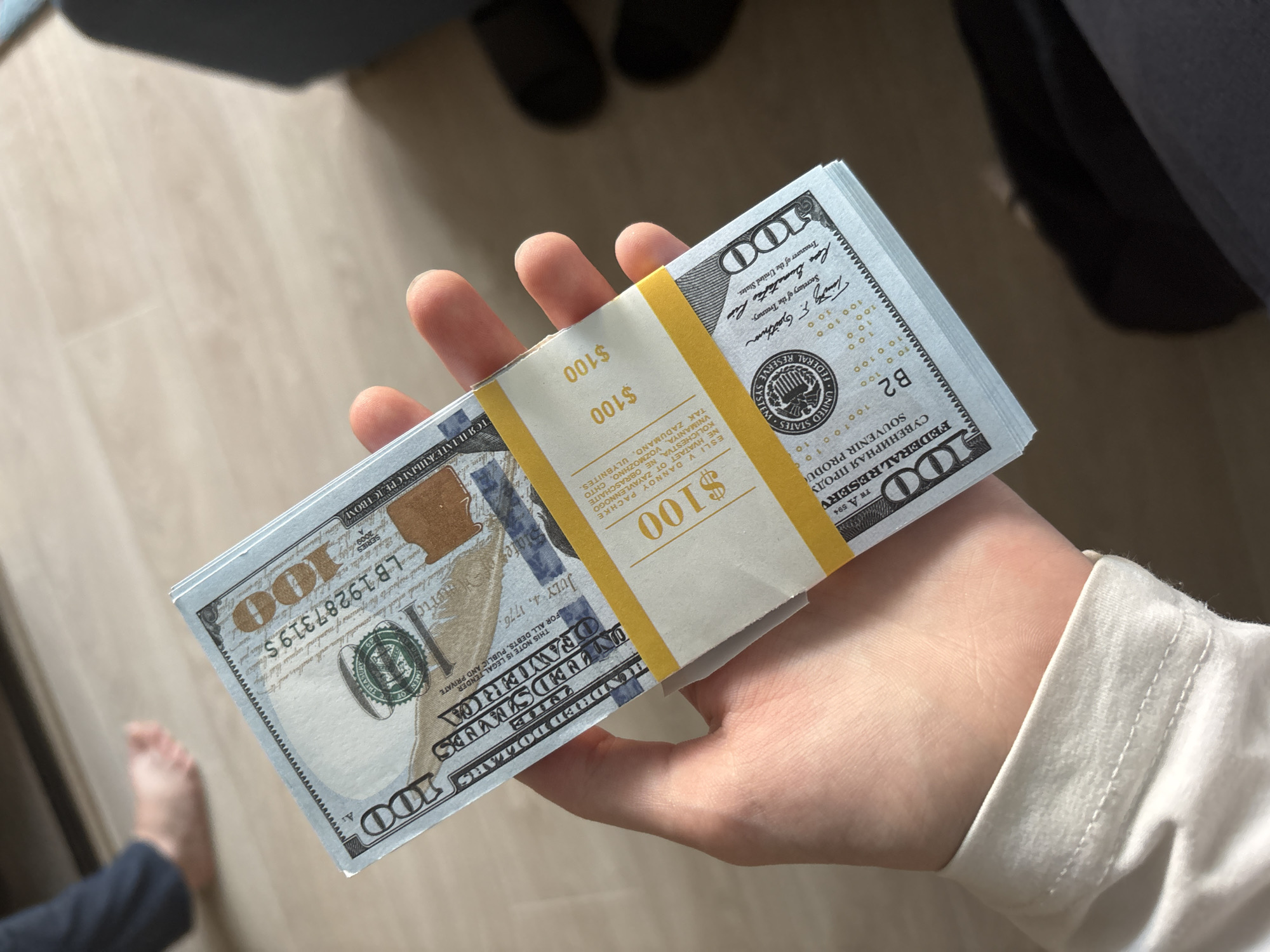 A person's hand holds a stack of U.S. $100 bills. The bills are banded together with yellow paper wrappers. The wrapper on the front of the stack is partially visible and has the text "$100" printed multiple times. The bills themselves have intricate designs, including portraits and text. The background shows a light-colored wooden floor and a blurred image of a bare foot and dark pants. The scene appears to be indoors.