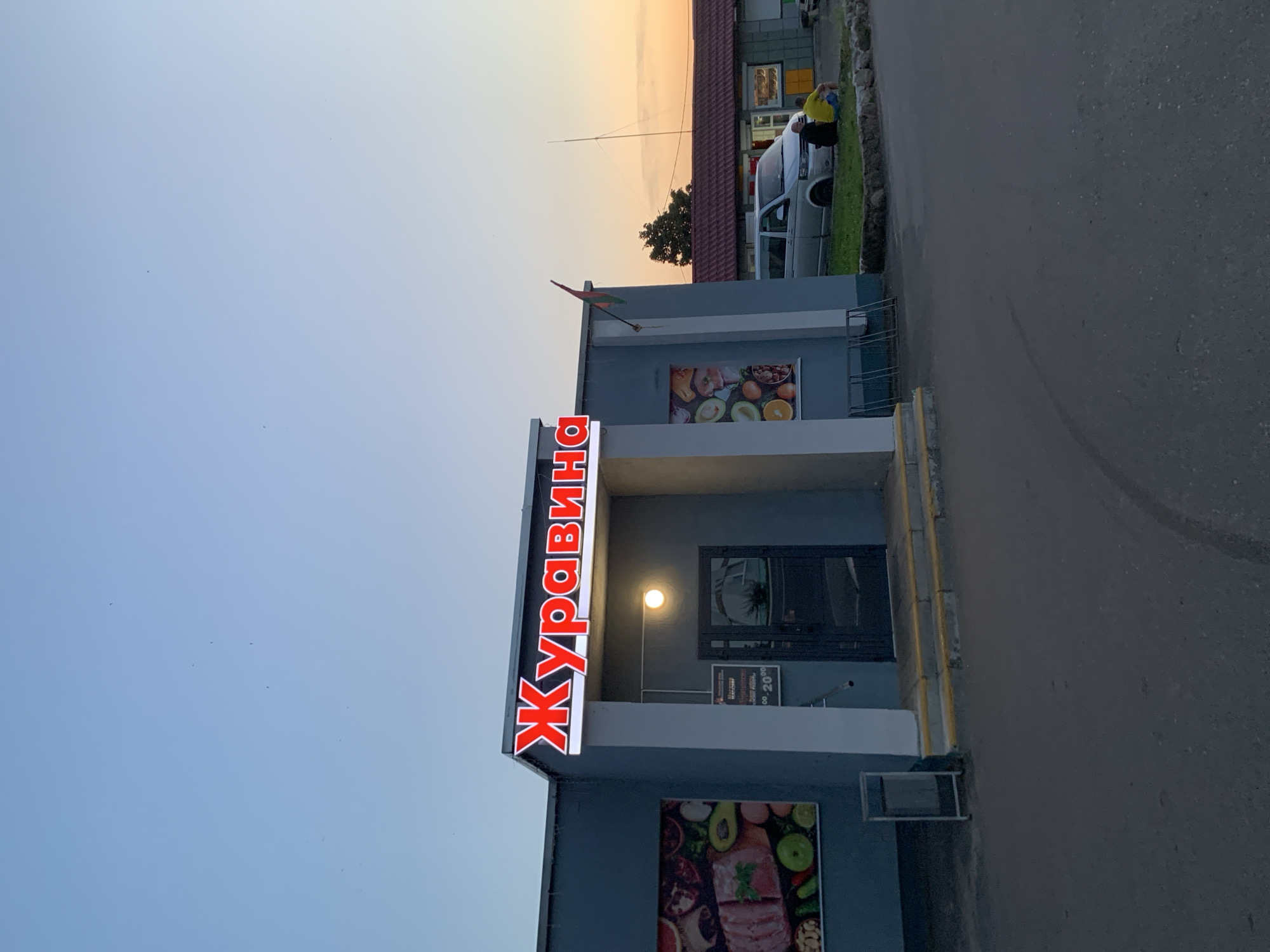 The image shows the front of a store in Kapyĺ, Belarus. A large, illuminated red sign with the word "ЖураВИНА" is mounted above the entrance. To the left of the entrance, there is a large poster featuring an assortment of fruits and meats. To the right of the entrance, a silver van is parked, and two individuals are seated on a grassy area near a building. A flag, possibly a national flag of Belarus, is visible on a pole attached to the store's exterior. The sky is clear and shows a gradient of light blue to orange, indicating it is either sunrise or sunset.