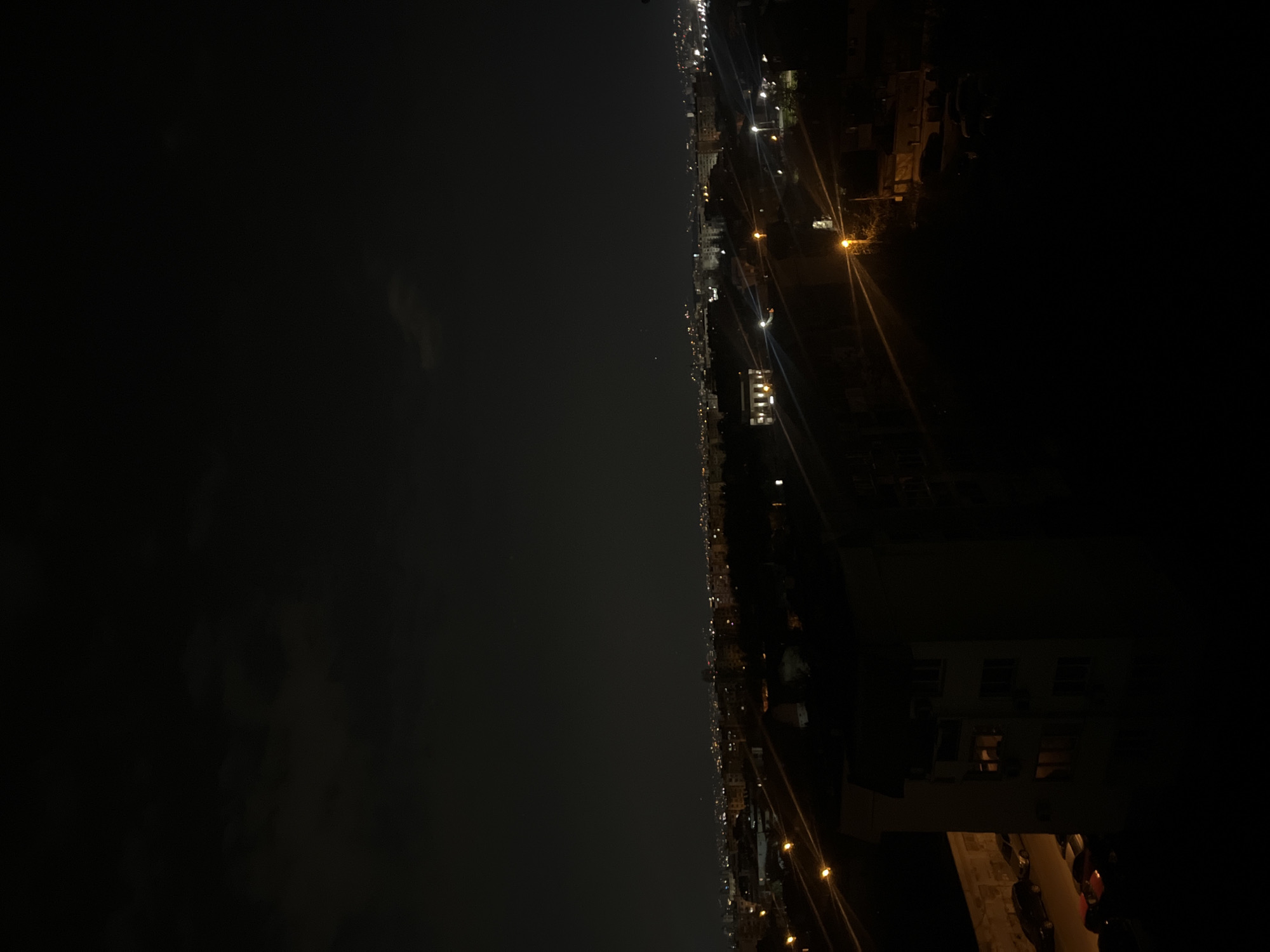 The image shows a nighttime urban scene, possibly in Sofia, Bulgaria, taken from an elevated position. The right side reveals a building with visible windows, parked cars, and streetlights. The city's skyline extends into the distance, with numerous lights indicating urban activity. The left side is largely dominated by a dark sky, likely reflecting the night sky over the city. Overall, the image captures a typical urban landscape at night.