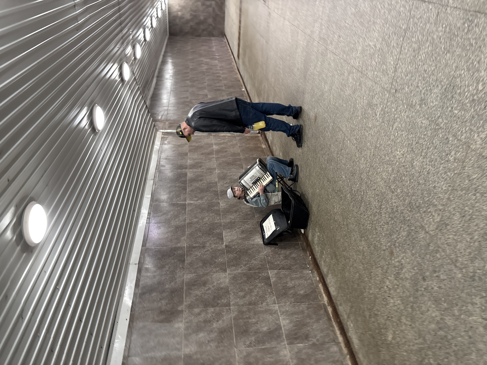 The image captures a scene within an underground pedestrian tunnel in Saint Petersburg, Russia. Two men are present, illuminated by a series of circular, flush-mounted lights along the left wall.
On the right side of the tunnel, a man is seated on a black foldable stool, playing a white and black accordion. He wears a light-colored baseball cap, a light-colored vest over a darker shirt, and blue jeans, appearing to be busking. Next to him on the floor is a black case, possibly for the accordion, serving as a music stand for sheet music. Visible text on the music includes "БЕЗУМНЫЕ ДЕБЮТАНТЫ" (Mad Debutants) and "ЧАСТУШКИ" (Chastushki), indicating the type of music being played.
A second man stands a short distance away, observing the musician. He is dressed in a black leather jacket, blue jeans, and dark shoes, wearing a yellow baseball cap and holding a yellow bottle or container. His posture suggests he is relaxed, possibly listening to the performance.
The tunnel features a light gray corrugated metal wall on the left, lined with bright circular lights. The right wall is a plain, textured light gray concrete surface. The floor is tiled with square, dark gray pavers. The artificial lighting suggests the time of day is either daylight, where the tunnel lacks natural light, or nighttime. The setting is functional and urban, typical of a pedestrian underpass.