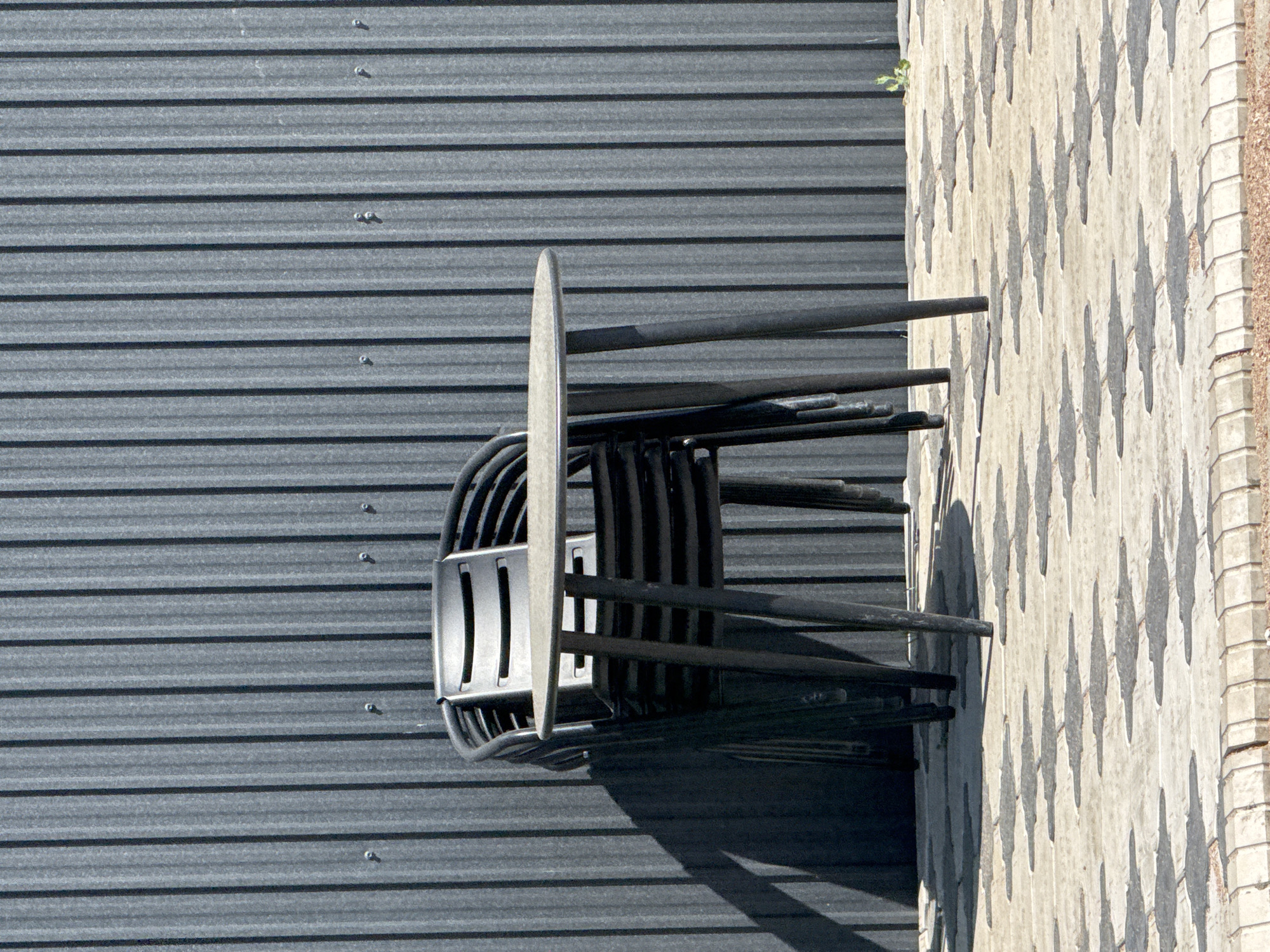 The image shows a stack of four black plastic chairs in front of a dark grey corrugated metal wall in the city of Sary-Oy, Kyrgyzstan. A small, round, grey table stands in front of the chairs, supported by three black legs. The scene is set on a light-colored paved patio with a patterned surface. Sunlight casts shadows from the table and chairs onto the patio. A small green plant grows near the right edge of the frame.