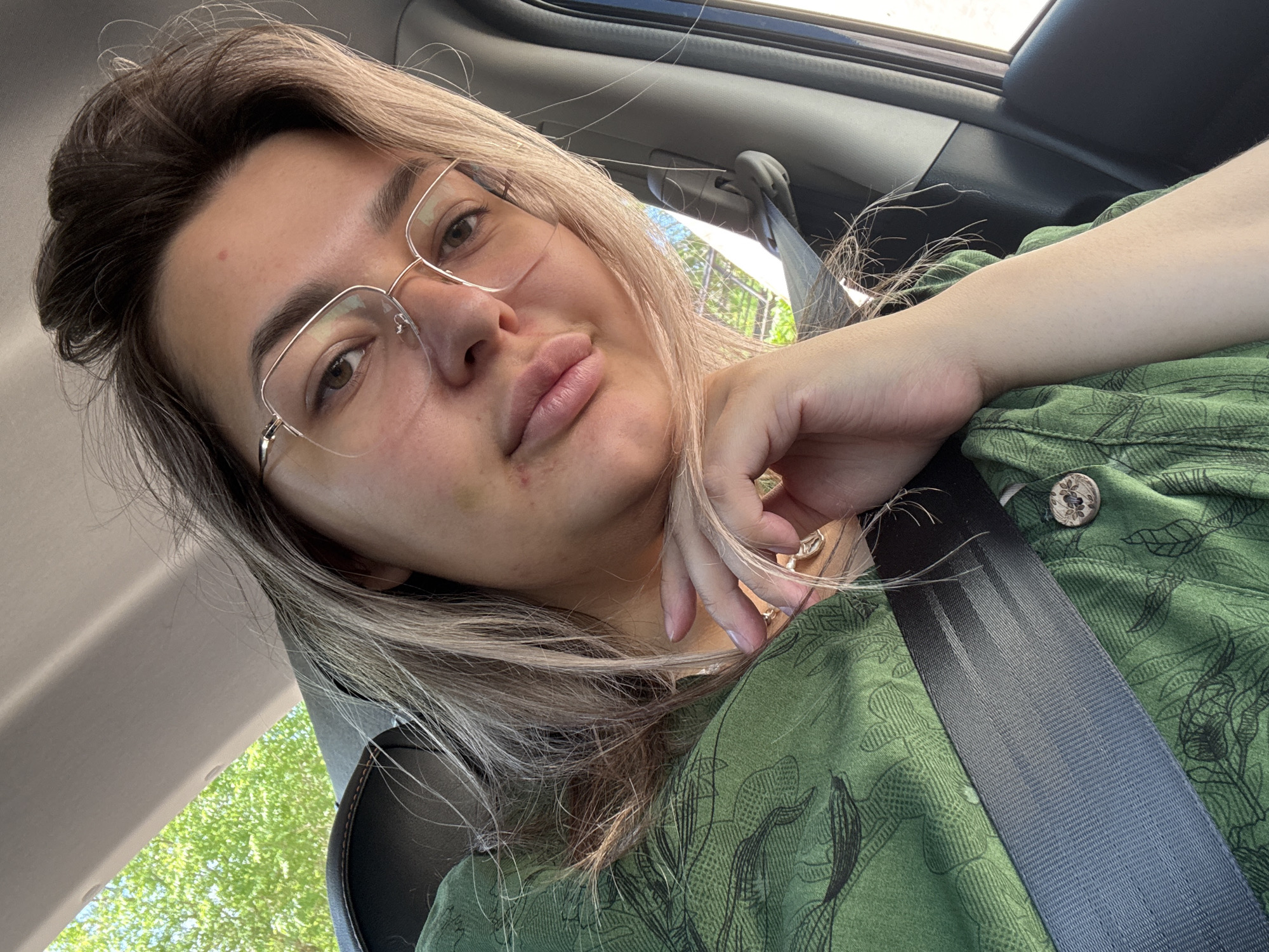 A woman is captured in a selfie from the chest up, inside a vehicle. She has light brown hair, some of which is falling forward, framing her face. She is wearing thin-rimmed glasses and a green top with a dark, leafy pattern and a wooden button. Her right hand is resting on her chin, and her left arm is visible, partially covered by her shirt. A seatbelt is fastened across her chest. The background shows a blurred view of green foliage, suggesting the vehicle is moving through a tree-lined area. The lighting suggests it is daytime, and the overall atmosphere is casual. This image was taken in the city of Birobidzhan, Russia.