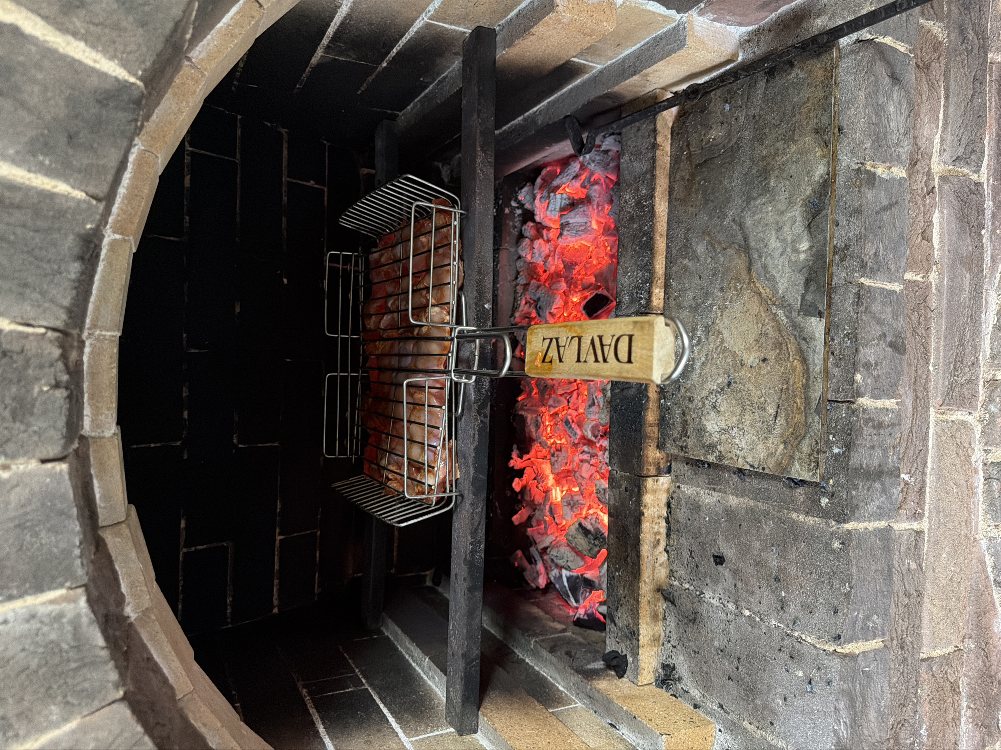 A close-up, eye-level shot captures a barbecue grill basket filled with raw sausages cooking over glowing red embers inside a brick oven. The grill basket is suspended from a wooden handle inscribed with the word "DAVLAZ". The oven's interior walls are made of dark brick, and the base is constructed from rough stones and lighter bricks. The overall setting appears to be an outdoor cooking area, possibly in a dacha or garden, consistent with the rural environment of Низинка Снт, Russia.