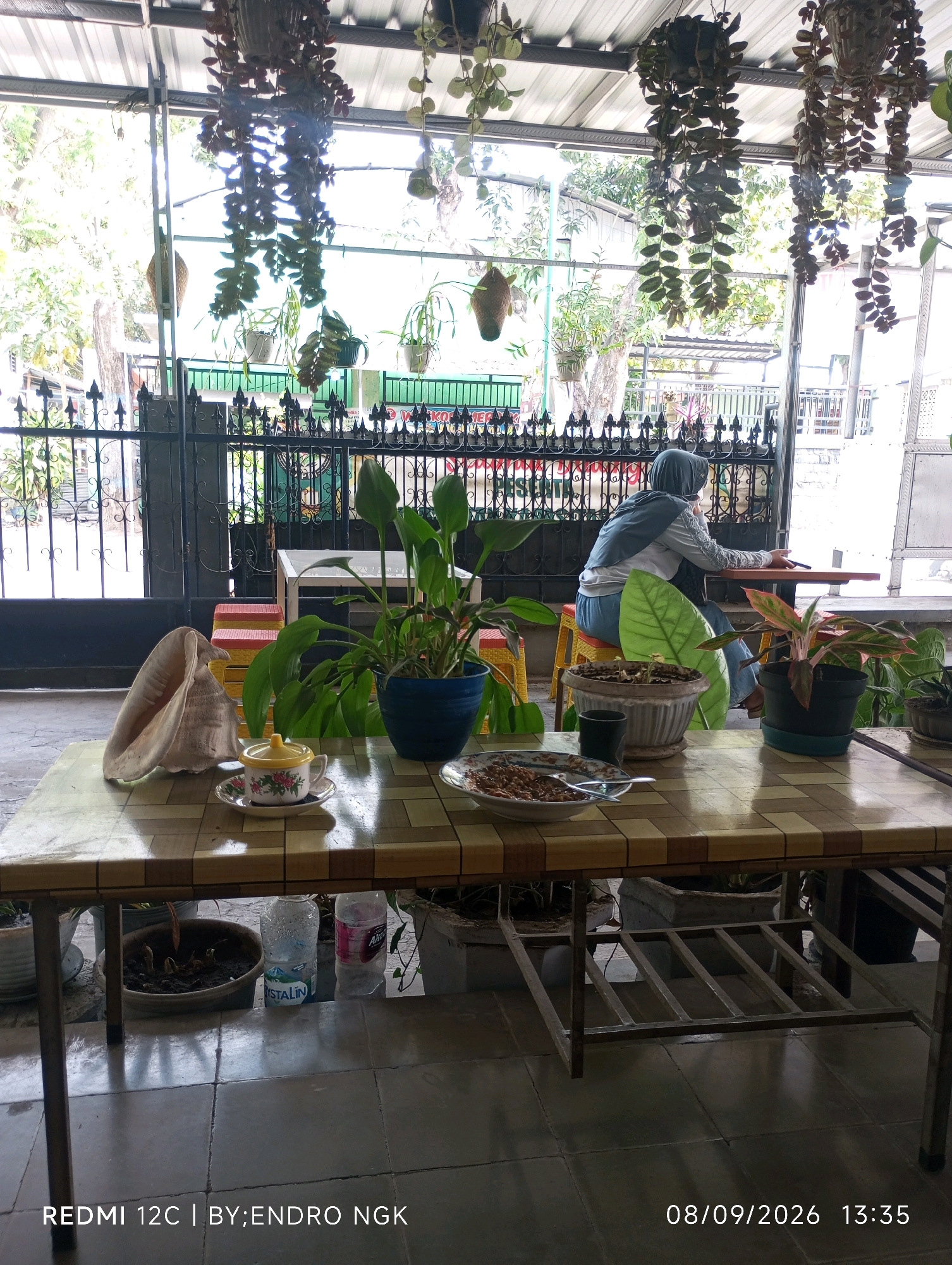 The image depicts an outdoor seating area, likely a cafe or restaurant in Nganjuk, Indonesia, based on the timestamp and model information. In the foreground, a rectangular table with a checkered top is set up on a tiled floor. Various items are placed on the table, including a large conch shell, a teacup and saucer with a yellow lid, a bowl of food, and a dark cylindrical object. Several potted plants are visible around the table and in the background, with some hanging from an overhead structure. A woman wearing a hijab and a light blue and grey outfit is seated at a smaller table in the background, looking towards the left and holding a smartphone. Behind her, a black metal fence encloses the area, and beyond that, trees and buildings are visible under a corrugated metal roof. The scene is lit by natural daylight.