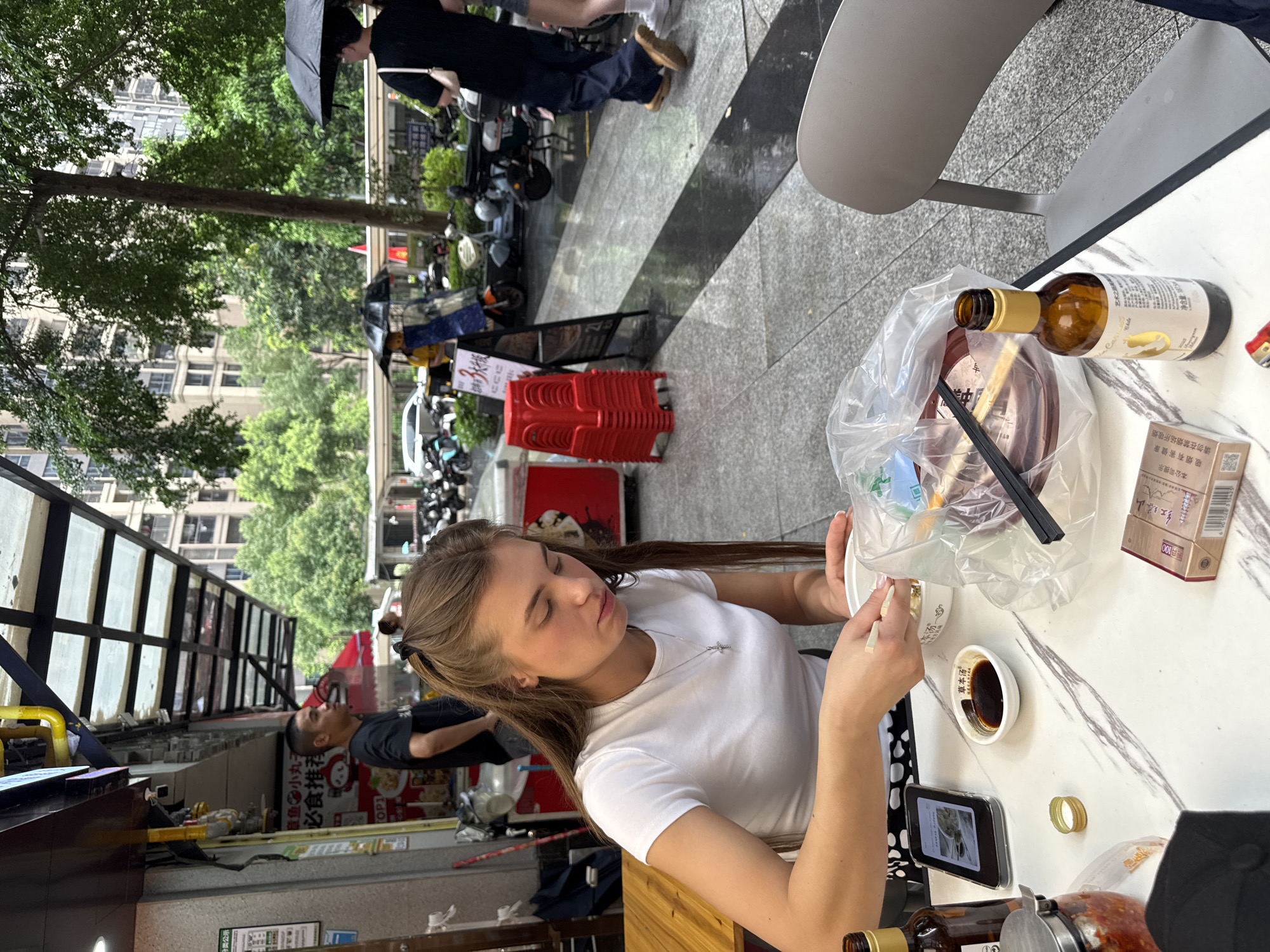 The image captures a street-level view of an outdoor dining area in Haizhu District, China. In the foreground, a young woman with long brown hair tied back is seated at a table, eating from a white bowl with chopsticks. She is wearing a white t-shirt and a silver cross necklace. On the table are several items, including a small bowl of soy sauce, a bottle of brown liquid, a smartphone, a container of what appears to be chili paste, a pack of cigarettes, and a plastic bag containing food. Behind her, a man in a black t-shirt stands near a food stall. In the background, more people are visible, some holding umbrellas due to light rain. Motorcycles and scooters are parked along the street. The environment suggests a bustling urban setting with covered walkways and apartment buildings in the distance. The ground is wet, indicating recent rainfall.