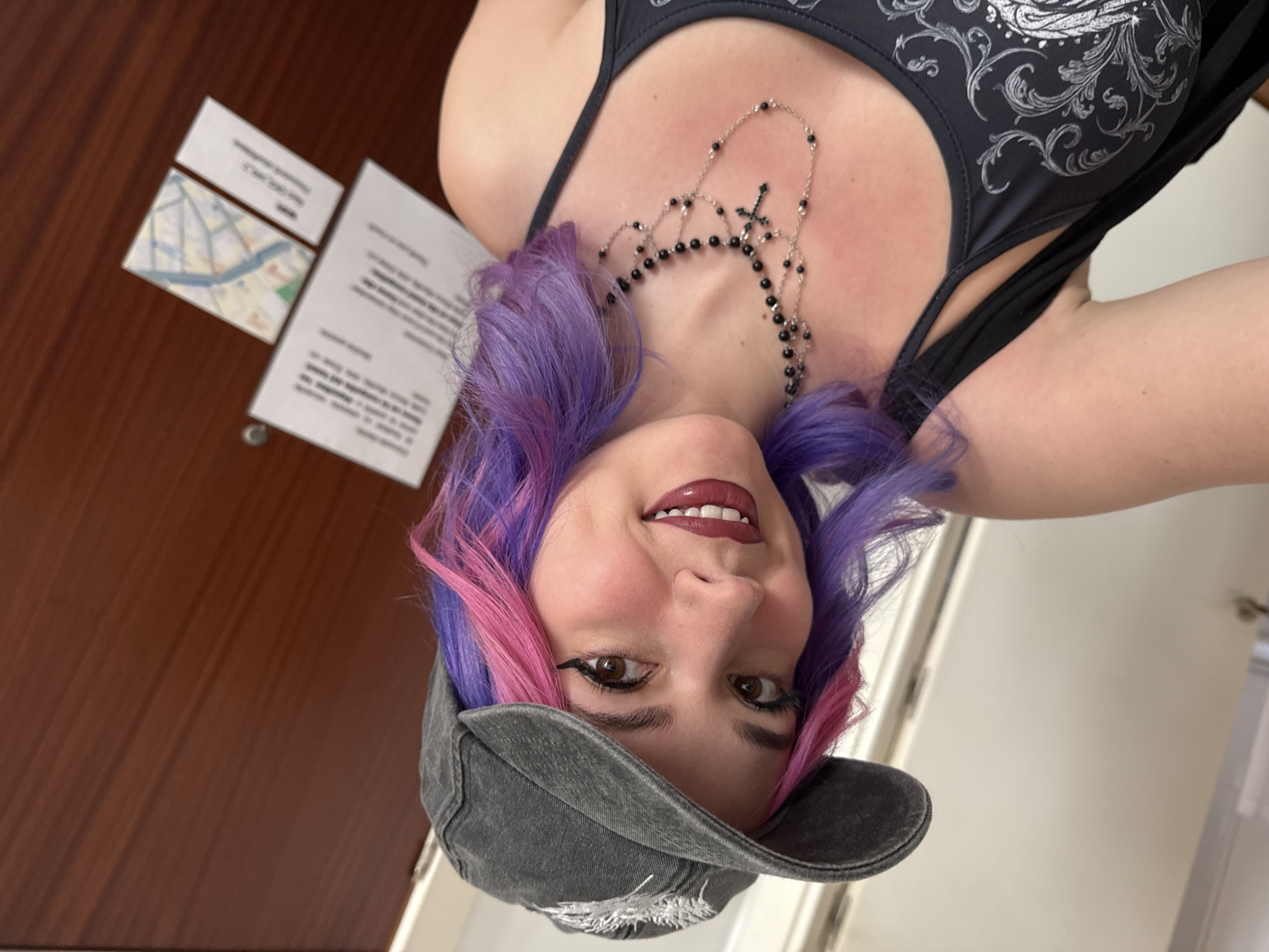 A person with dyed pink and purple hair is wearing a black tank top with a silver ornamental design and a black beaded necklace with crosses. They are wearing a grey baseball cap and dark lipstick. To the left is a brown wooden door with two white informational pamphlets attached, one showing a map of a section of Madrid.