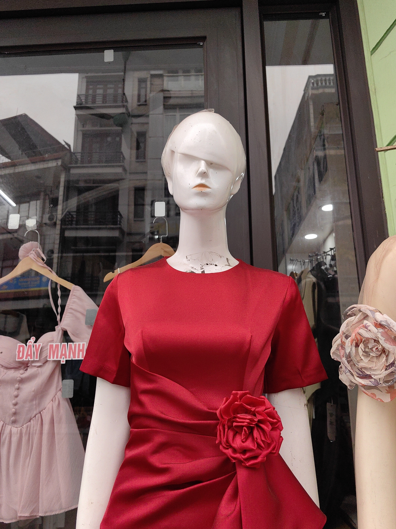 A red dress is displayed on a mannequin in front of a window in Bai Chay Ward, Vietnam. The window reflects the exterior buildings and a pink dress is visible on a hanger.