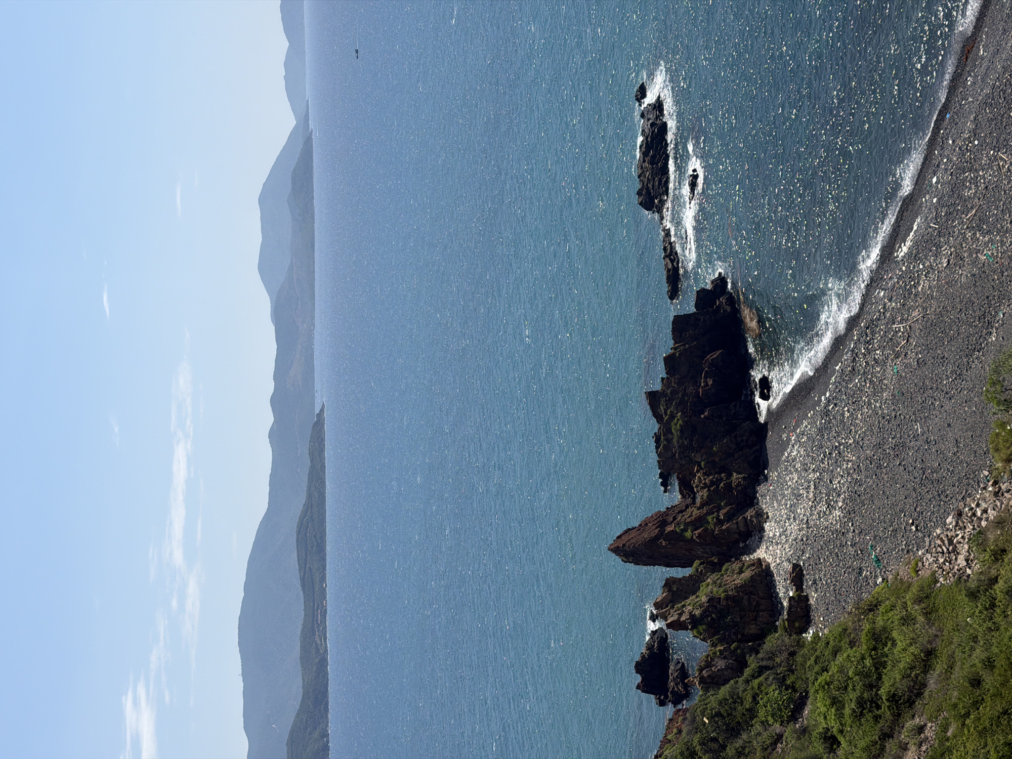 This media file captures a serene coastal landscape in Nam Nha Trang Ward, Vietnam, under a bright, clear sky. The image features a vast expanse of shimmering dark blue ocean, reflecting sunlight and creating numerous bright glints on its surface.
On the left side of the frame, a series of hazy, dark blue-green mountains recede into the distance, suggesting a rugged coastline or island chain. Far out on the water, a small, dark, indistinct object, possibly a boat, can be seen against the horizon.
The right side of the image is dominated by a dramatic, rocky coastline. Jagged, dark brown and black rock formations jut into the water, with white foamy waves crashing around their bases. Adjoining these rocks is a crescent-shaped beach composed of dark pebbles or gravel, where gentle white waves meet the shore. The immediate foreground on the right features lush green vegetation and bushes, growing on the land leading down to the rocks and beach.
The time of day appears to be late morning or early afternoon, characterized by bright sunlight and minimal cloud cover, with only a few wispy clouds visible in the sky. There is no visible text, nor are any people or animals explicitly discernible within the scene. The overall impression is one of natural beauty and tranquility.