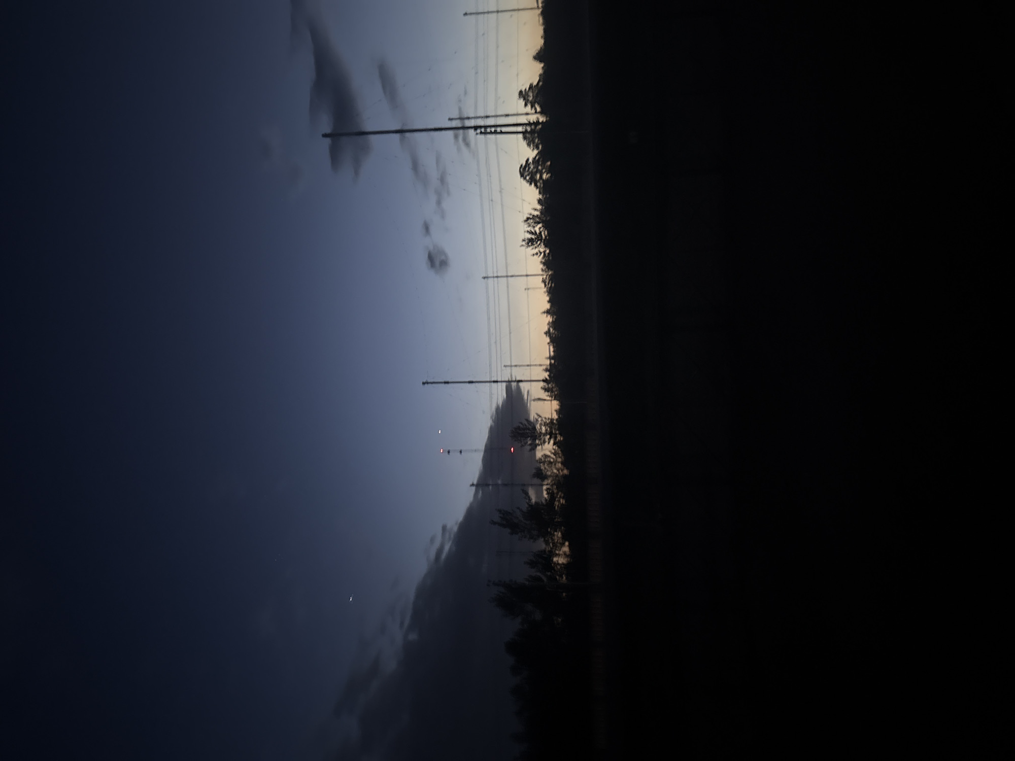 The image shows a view at dusk of an urban edge, with electricity pylons rising against a cloudy sky. The city is potentially located in Unknown, Unknown. The area is bordered by dark vegetation, which could represent a park or natural area. The pylons suggest the presence of infrastructure, possibly indicating an industrial or suburban fringe of the city. No people are visible. The environment is transitioning from daylight to nighttime.