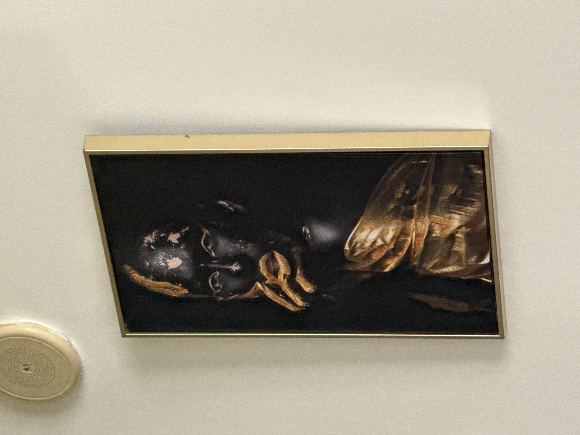 A framed canvas print featuring a portrait of a person with black body paint and gold accents is displayed on a textured off-white wall. The print is oriented vertically and has a thin gold frame. In the upper left corner of the image, a cream-colored circular object with perforations is visible, attached to the wall. This object appears to be a smoke detector or a speaker. The scene is indoors. There is no indication that this scene is located in Dubai, United Arab Emirates.