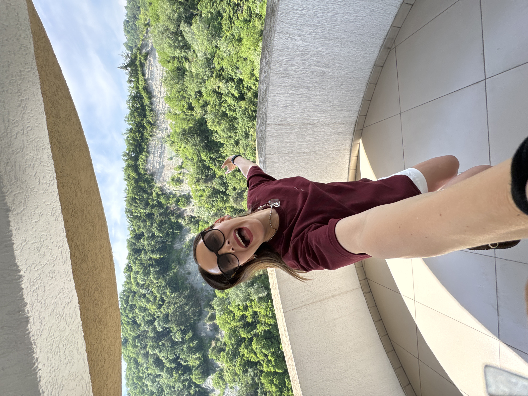 A woman is taking a selfie on a balcony overlooking a lush, green mountainside in Sochi, Russia. She is wearing sunglasses, a maroon t-shirt, and white shorts. Her mouth is open in a laugh or shout, and she is pointing with her left hand. The balcony has a curved white railing and a tiled floor. The background is dominated by dense green trees covering the slopes of a mountain, with layers of rock visible on the mountainside. The sky is blue with scattered white clouds. The overall impression is one of joy and enjoyment of the natural scenery.
