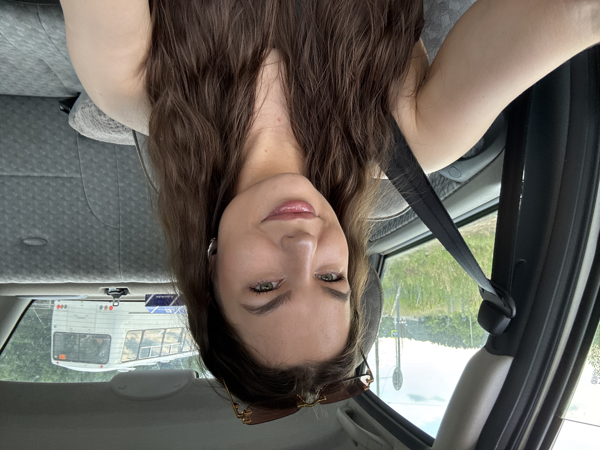 A young woman with long, wavy brown hair is taking a selfie inside a car. She is wearing sunglasses on top of her head and has hoop earrings in her ears. The woman is smiling, and her lips are glossy. She is wearing a dark-colored top. The car's seatbelt is visible across her shoulder. Outside the car window, there is greenery and a signpost. In the distance, a white bus is visible. The background appears to be a semi-urban or rural area.