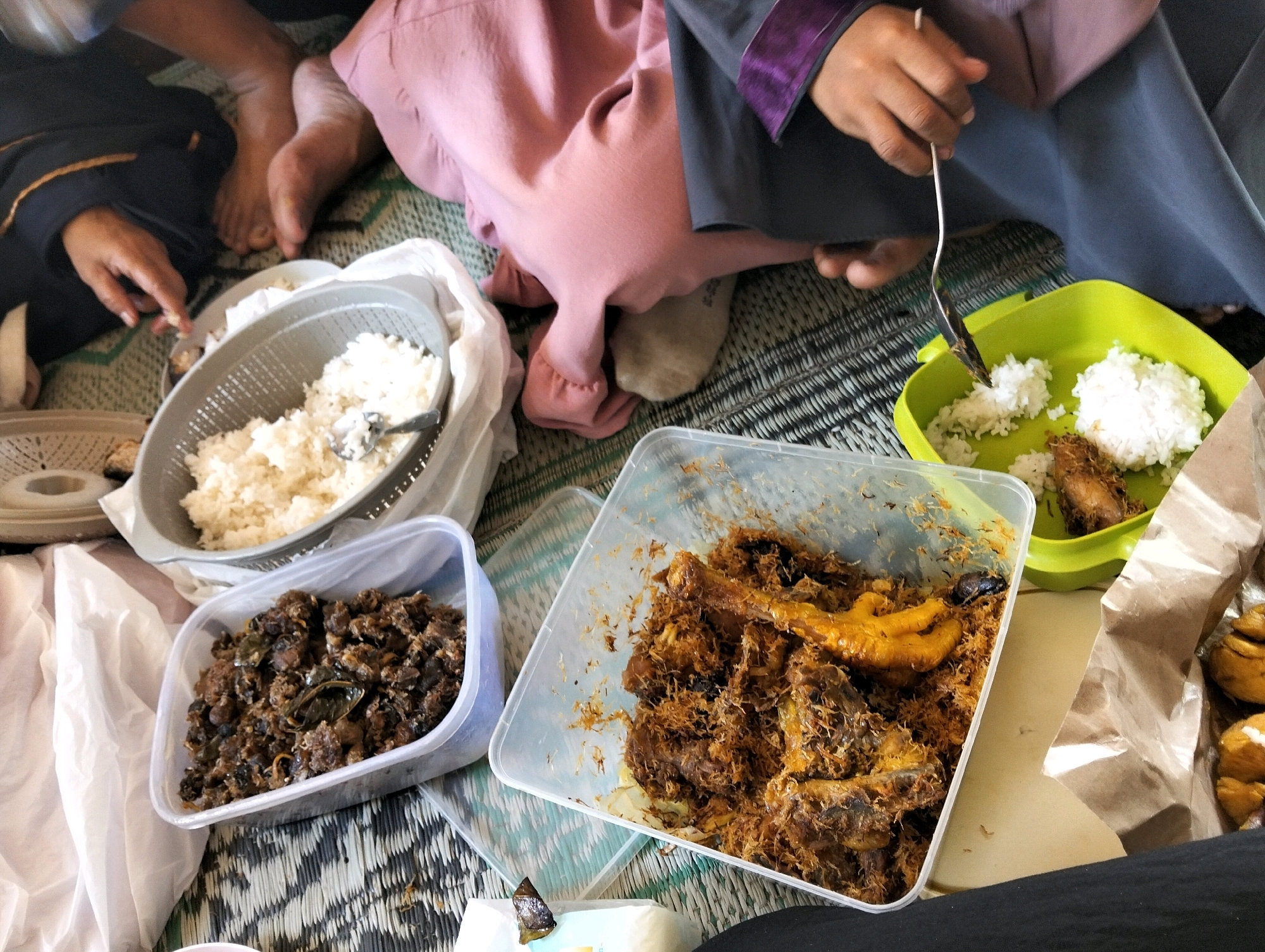 In Subang, Indonesia, a group of people are having a meal outdoors, seated on a woven mat. The scene focuses on a variety of dishes laid out for consumption. In the foreground, a translucent plastic container holds a brown, finely shredded meat dish, possibly a traditional Indonesian delicacy. To its right, a larger clear plastic container is filled with a more substantial meal, featuring pieces of chicken and what appears to be a chicken foot, all coated in a similar shredded, brown seasoning. A bright green plastic plate contains white rice and a piece of fried chicken. To the left, a grey plastic colander overflows with cooked white rice, and a spoon is partially submerged in it. Another smaller, out-of-focus container is visible to the far left. The people are dressed in casual attire, with one person wearing a pink garment and another a grey robe with purple trim. Their legs are visible, some bare, suggesting a relaxed, informal setting. The overall impression is of a communal meal enjoyed in a warm, outdoor environment.