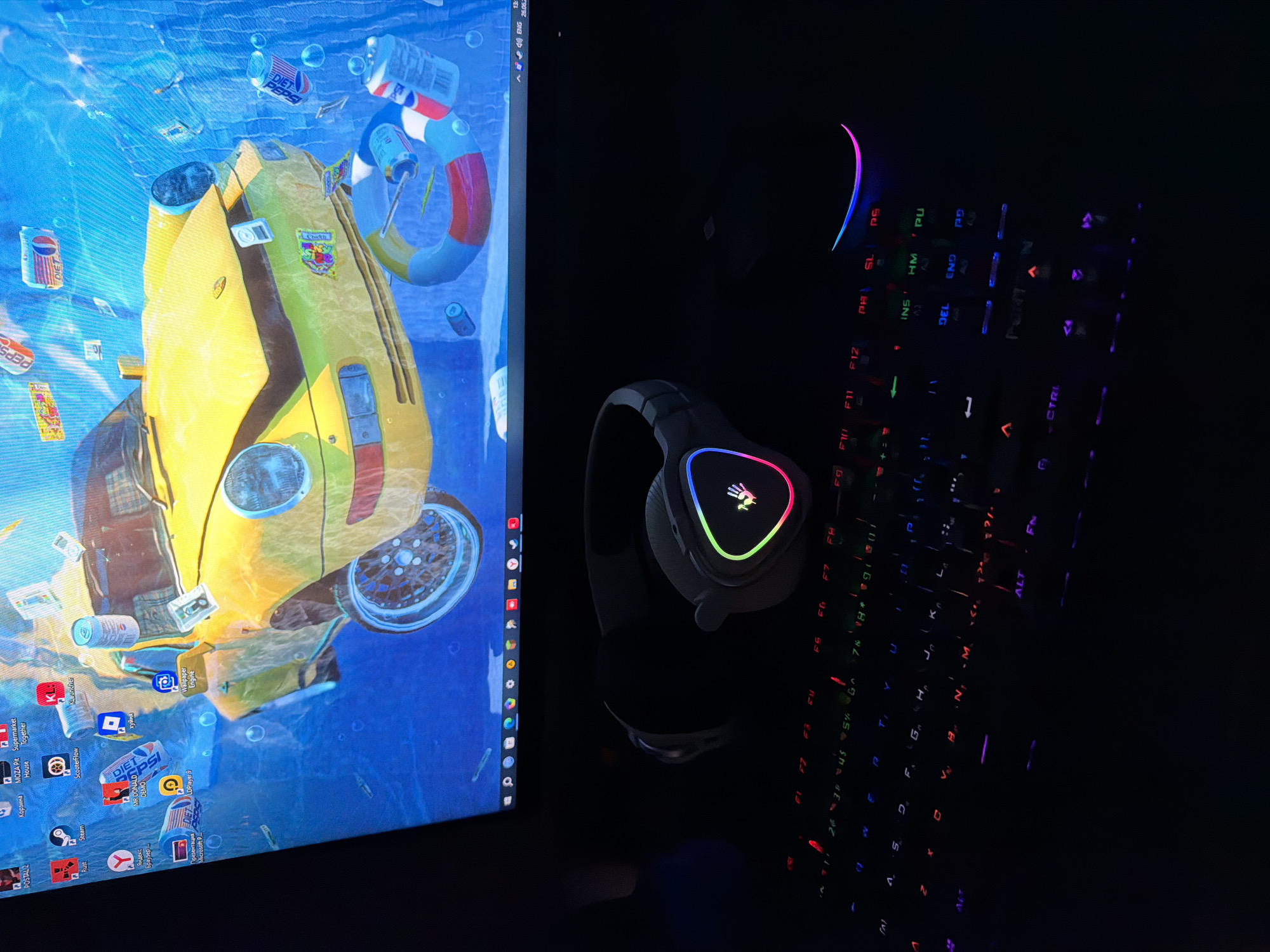 The image displays a computer setup with a desktop monitor showing a vibrant wallpaper of a yellow car submerged underwater, surrounded by Pepsi cans and money. The monitor also shows taskbar icons including icons for "POSTAL 2", "Коронавирус", "MOZAIKA", "Supermarket", "Rust", "Steam", "ScooterFlow", "Klouncher", "Mr. DONALD DEMO", "Roblox", "Wallpaper Engine", "Yandex Browser", "Microsoft PowerPoint", and "LDPPlayers". Below the monitor, a gaming headset with a glowing logo is placed on a dark surface, likely a desk. A black keyboard with illuminated keys, displaying a variety of colors, is also visible in the foreground. The scene is dimly lit, with the keyboard and headset lights being the primary light sources in the lower portion of the image. The image is taken from a slightly high angle looking down at the keyboard and headset. No specific location in Krasnoyarsk, Russia, can be identified from the image itself.