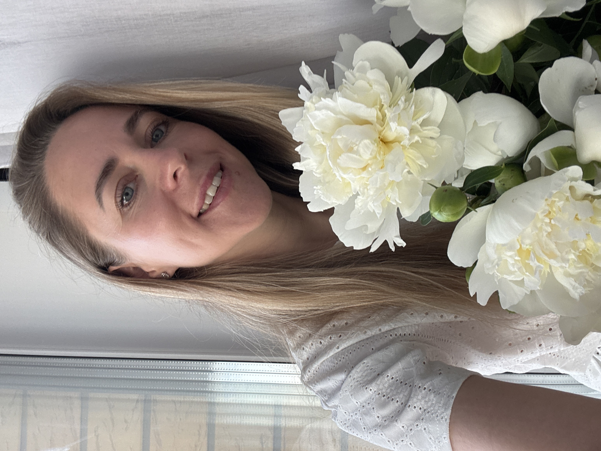 A blonde woman is pictured at eye level, holding a bouquet of white peonies. She is smiling, looking towards the camera, with her head tilted slightly. The woman has blue eyes and fair skin, and is wearing a white eyelet blouse. She is positioned in front of a window with vertical blinds on the left side of the frame, and a white curtain on the right. The background is a plain wall. The lighting is natural, coming from the window. The image appears to be a selfie taken indoors, possibly in an apartment in the city of Chaikovsky, Russia, given the context.