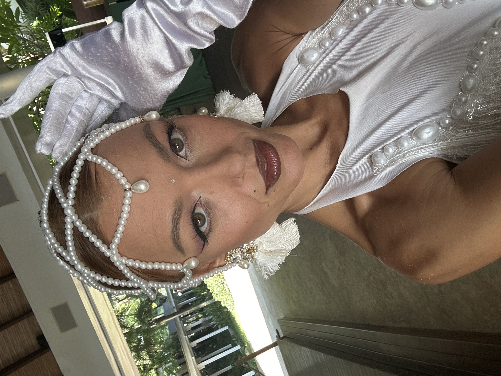 In Ko Samui District, Thailand, a woman dressed in a white costume adorned with pearls and sequins poses for a selfie. She wears an elaborate pearl headdress and large tassel earrings. Her right hand, covered in a white satin glove, is raised to her forehead. The background suggests an outdoor setting with a wooden-tiled roof and greenery visible. The woman's makeup is dramatic, with winged eyeliner and dark lipstick.