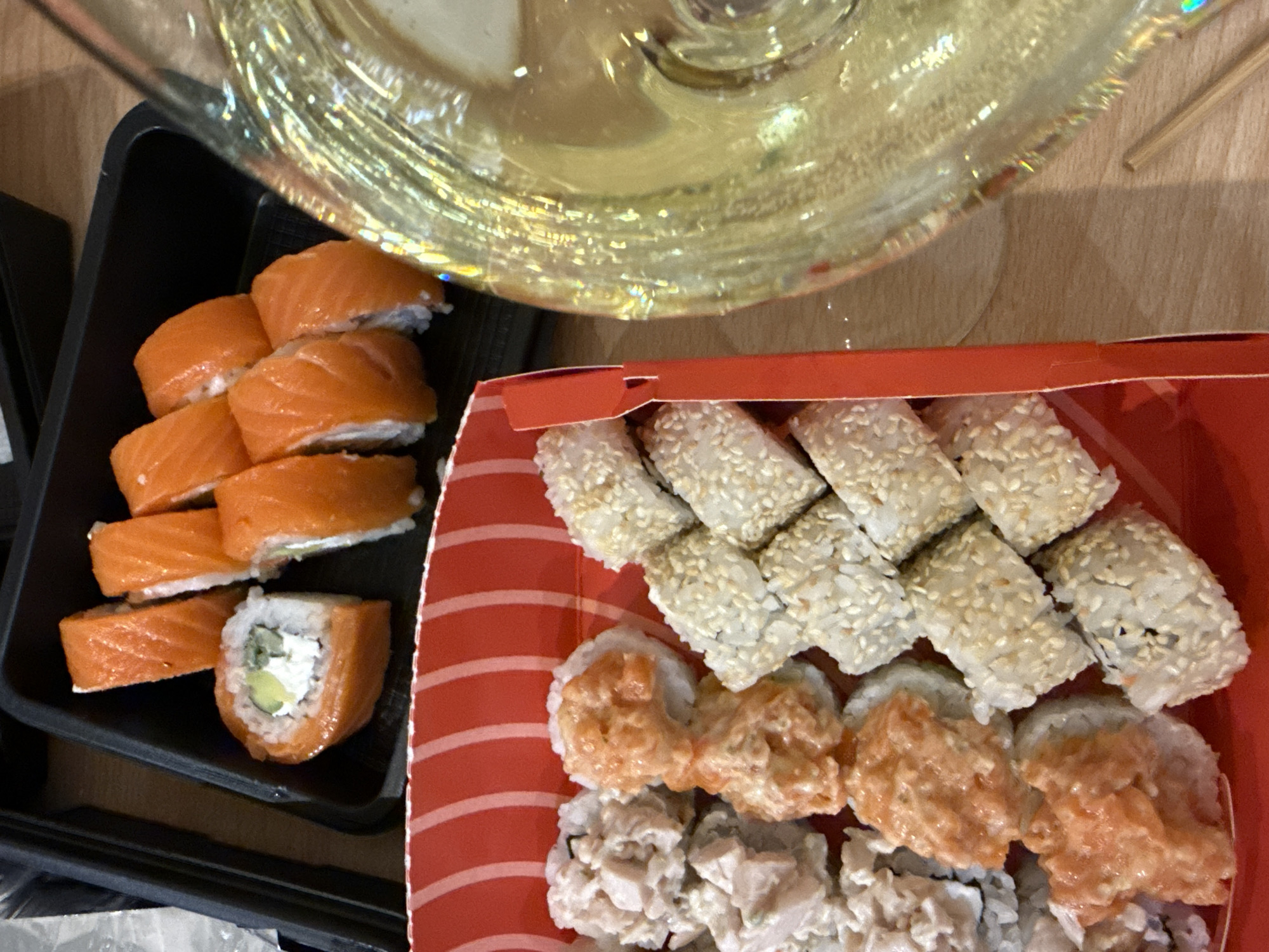 The image shows two containers of sushi arranged on a light brown surface, possibly a table. The container on the left is black and contains approximately 10 pieces of salmon sushi, some with visible avocado and cream cheese filling. The container on the right is red and contains three rows of sushi rolls: one row covered in sesame seeds, one with salmon topping, and another with a shredded white topping. A glass, possibly containing white wine, is also partially visible in the upper portion of the image. The scene appears to be indoors. There is no indication of the location being Kazan, Russia, or any information about people or interactions.