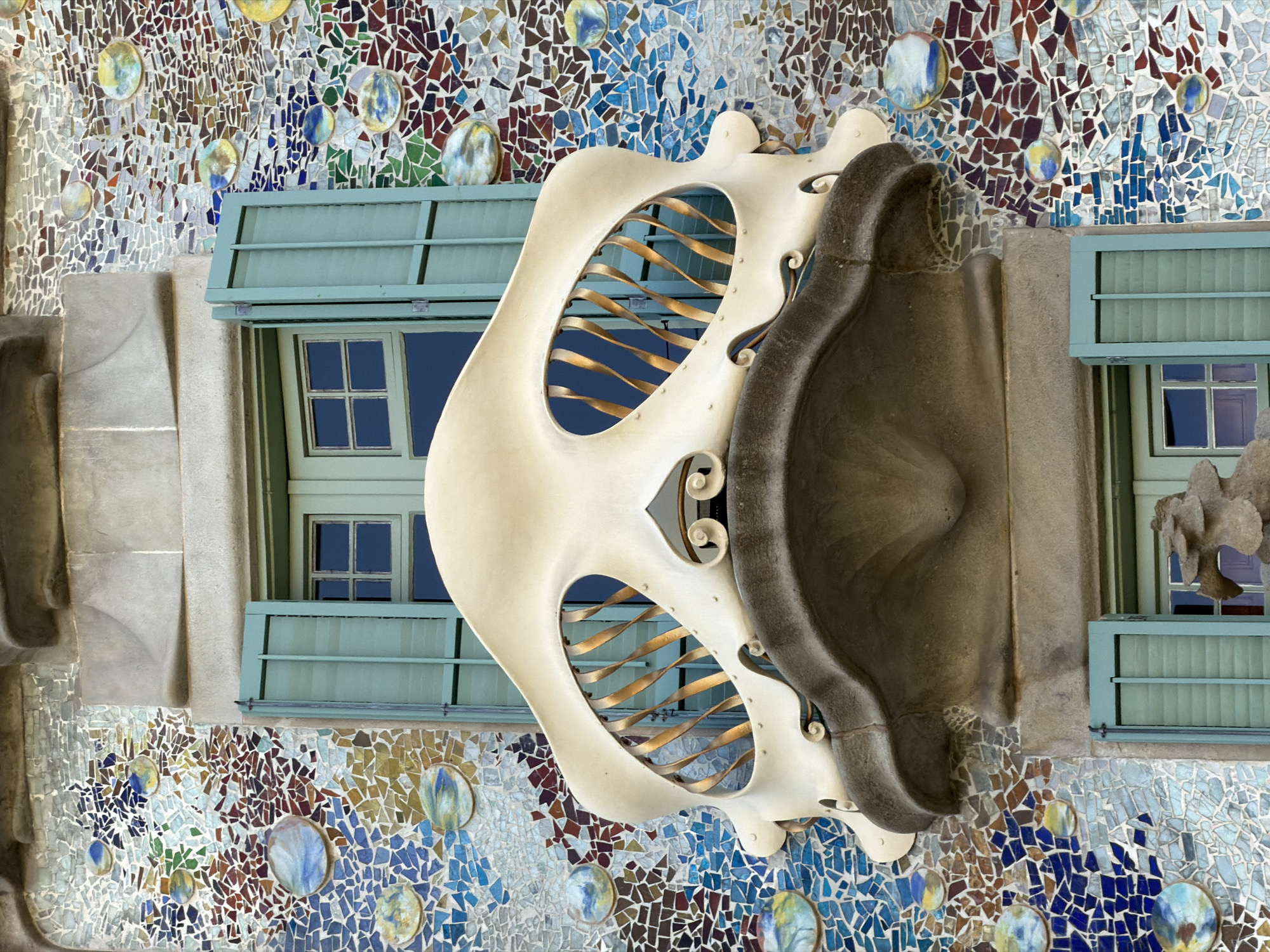 The image shows a close-up view of a balcony on Casa Batlló, a building designed by Antoni Gaudí located on Passeig de Gràcia in Barcelona, Spain. The balcony is a distinctive organic shape, appearing almost like a large, stylized bone or a mouth. It is made of cream-colored material and features gold, twisted metal railings visible through an oval opening. The wall surrounding the balcony is covered in a mosaic of colorful, irregular tiles in shades of blue, white, red, green, and brown, interspersed with circular ceramic elements that resemble bubbles or planets. A light blue shuttered window is visible behind the balcony, and another window with a similar shutter is seen below. The architectural style is characteristic of Gaudí's Art Nouveau designs, known for its flowing lines and vibrant, nature-inspired ornamentation. There are no people visible in the image.