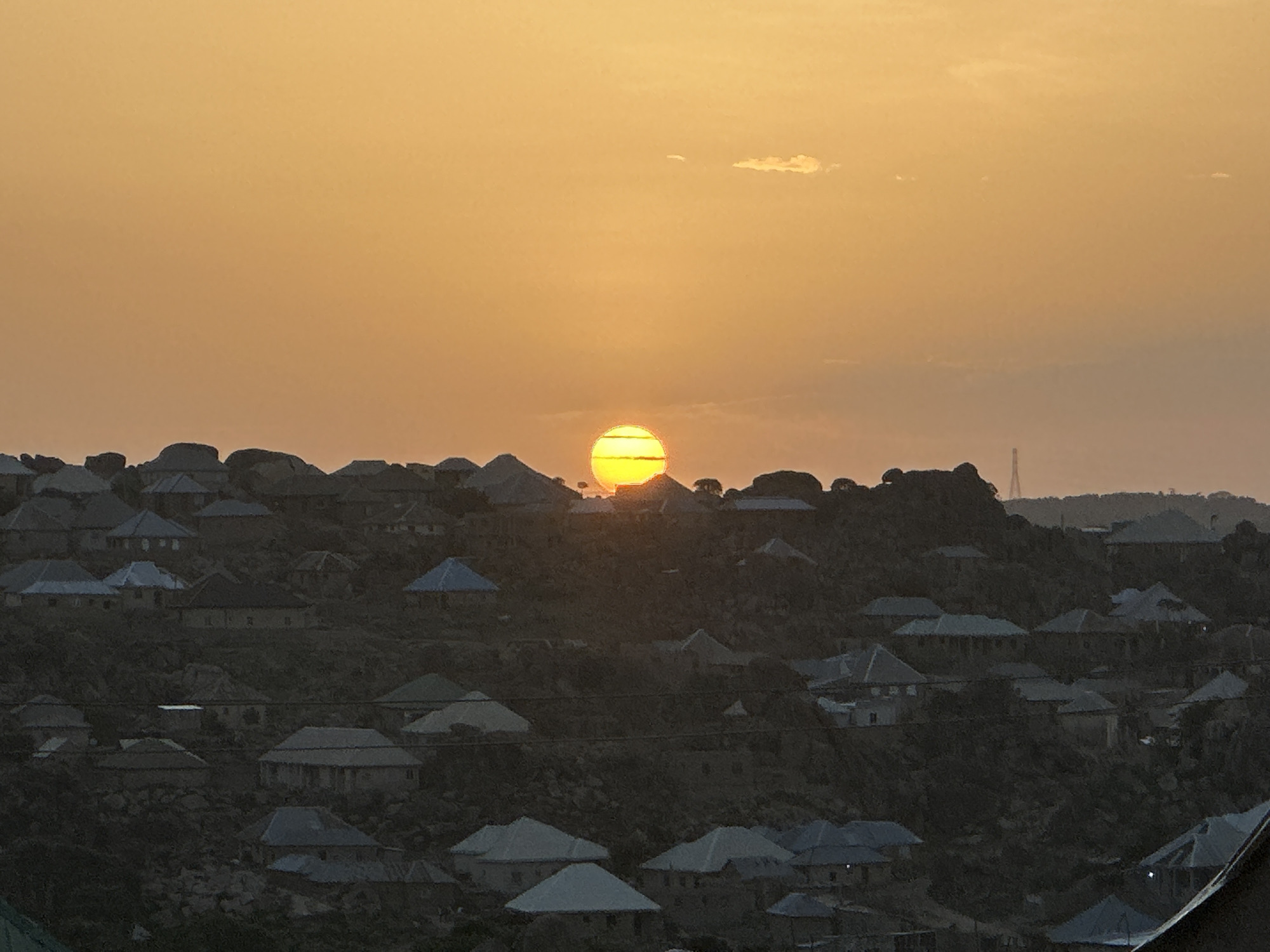 The image shows a sunset over a densely populated hillside in Jos, Nigeria. Numerous houses with gray and blue roofs are visible, built on the slope. The sun is low on the horizon, partially obscured by the hill's crest, casting an orange glow across the sky. A distant structure resembling a tower is visible on the horizon.
