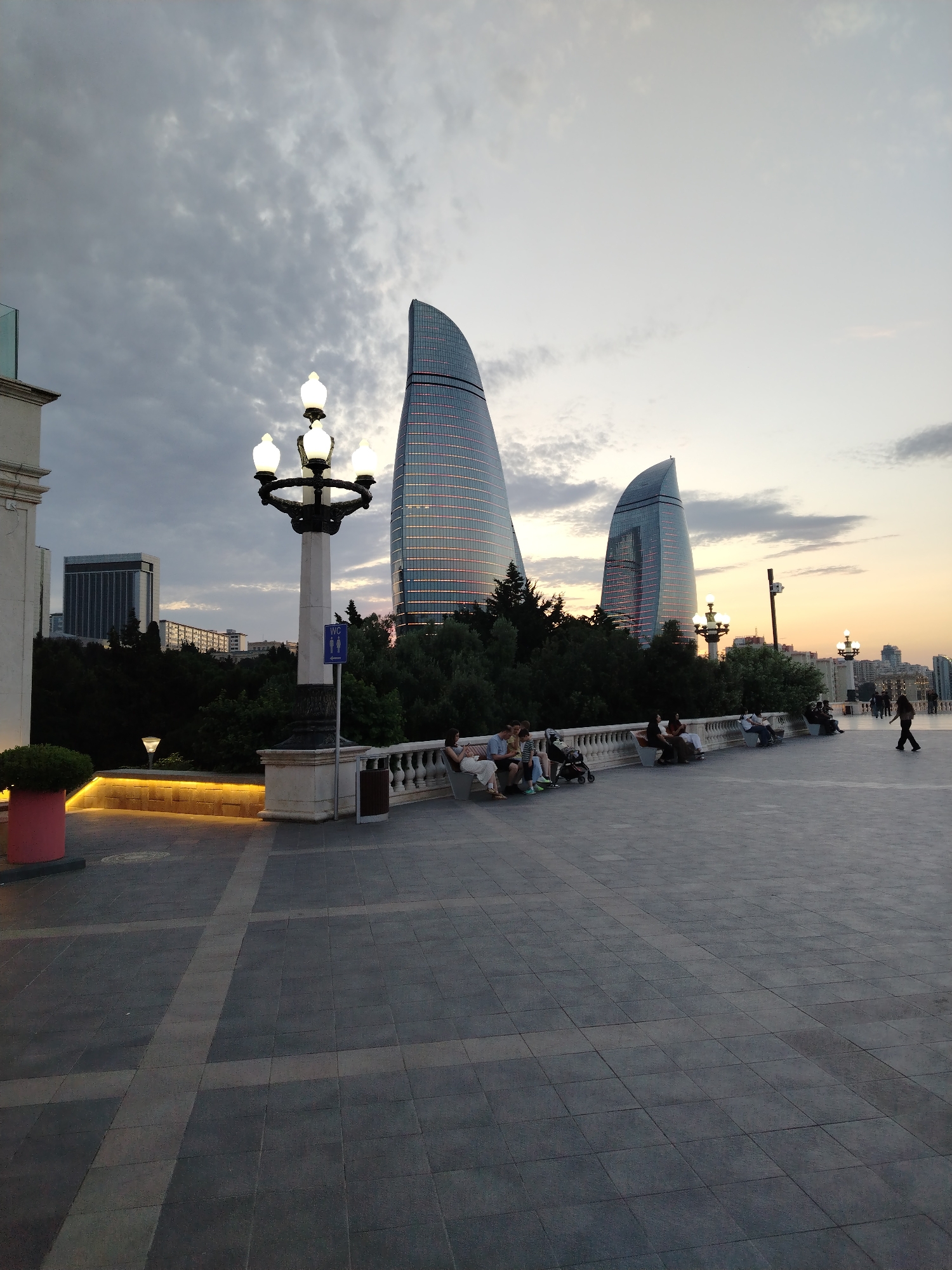 The image shows a wide outdoor plaza paved with dark gray square tiles. The plaza is lined with white ornate railings. Several people are seated on benches along the railings, observing the scenery. In the background, two distinctive flame-shaped skyscrapers dominate the skyline, their glass facades reflecting the dimming twilight sky. The sky is filled with dramatic gray clouds, hinting at dusk or an overcast evening. A large lamppost with multiple lights stands prominently on the left side of the plaza, near a sign with male and female figures, indicating a restroom. To the left of the lamppost, a white building with a glass railing is visible. Lush green trees form a dense line behind the railing. In the foreground, a portion of a pink planter with a green shrub can be seen. The overall scene suggests a public space in Baku, Azerbaijan, likely the Boulevard or a similar scenic viewpoint overlooking the city and its iconic architecture.
